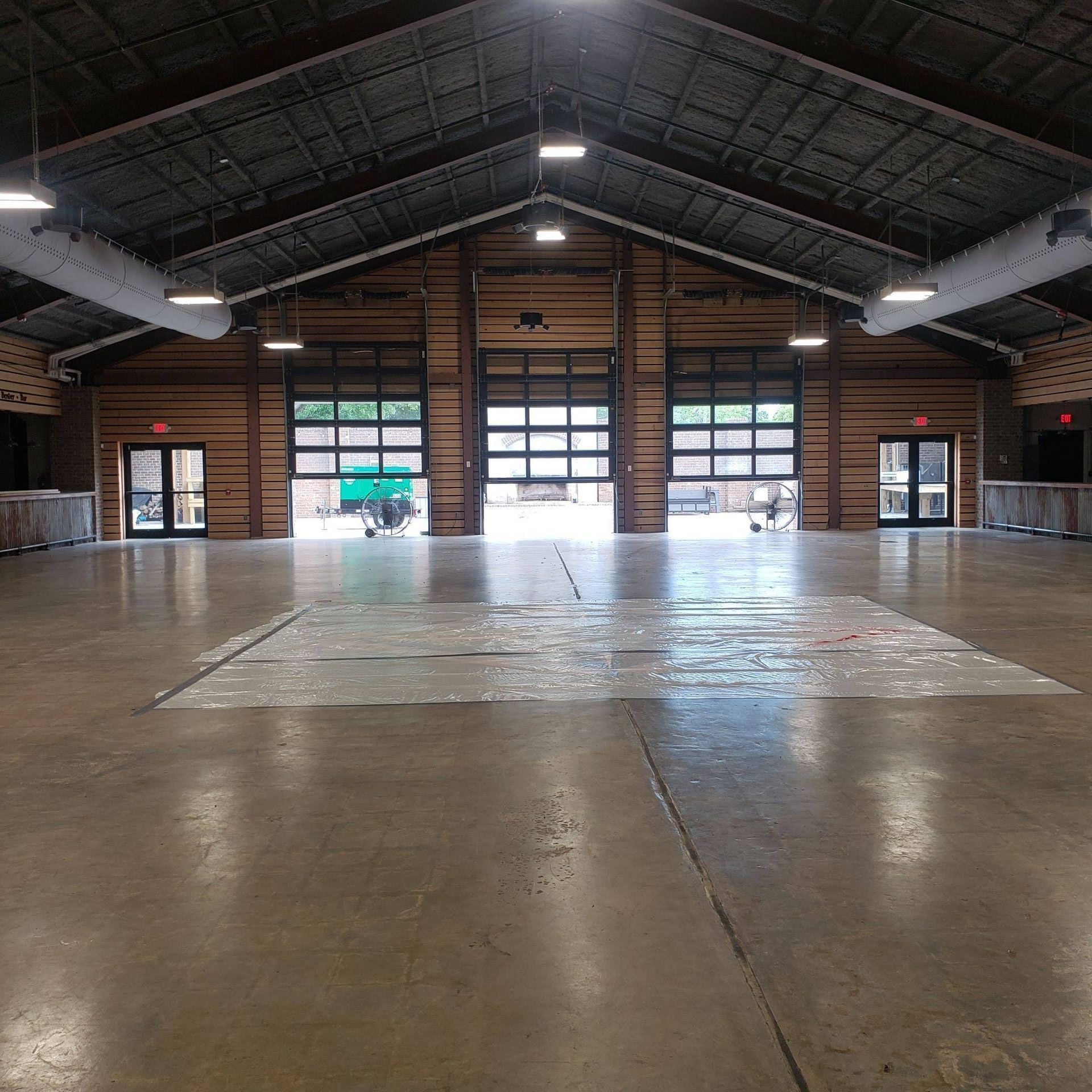 A large, empty event hall with high ceilings, wood-paneled walls, a polished concrete floor, and multiple garage doors.