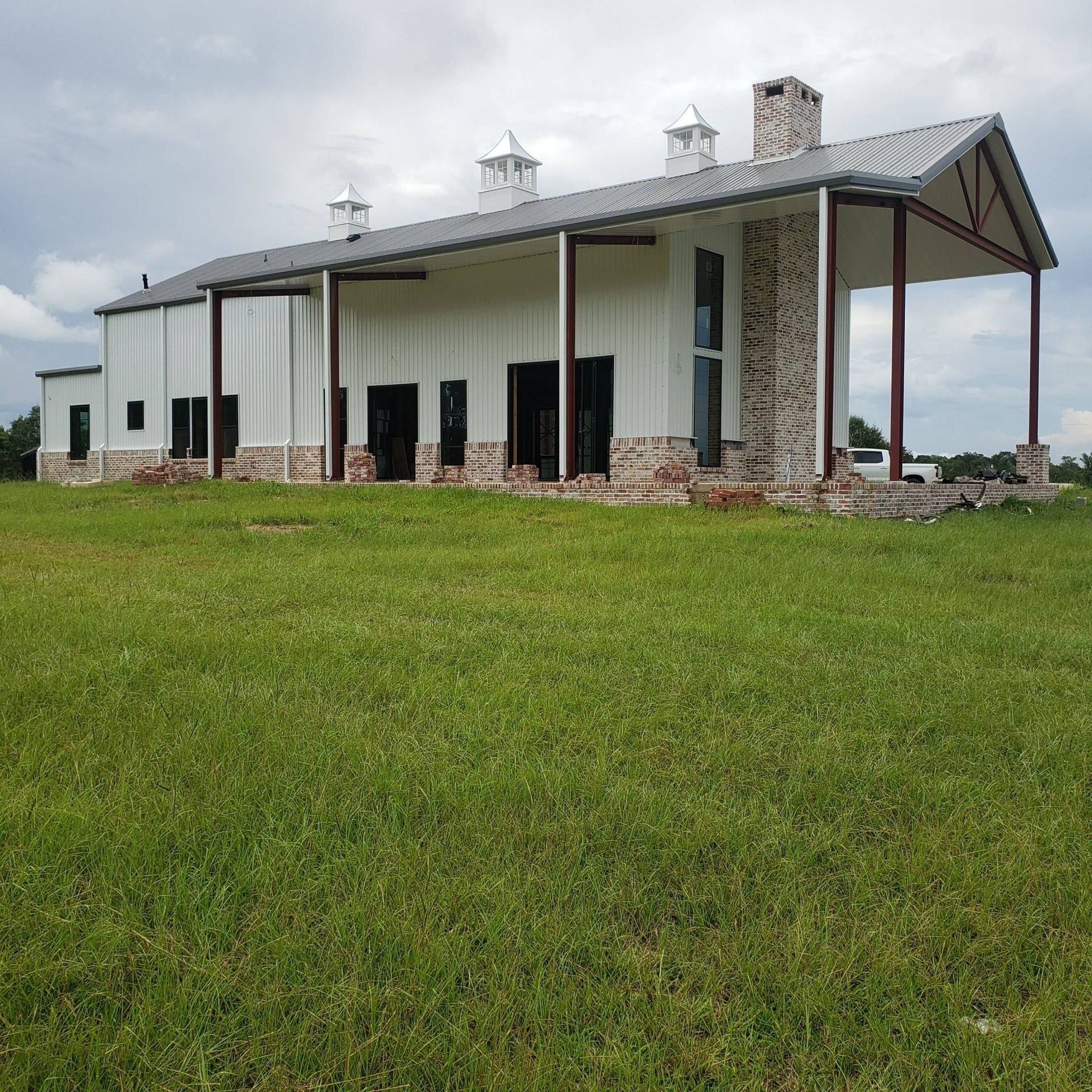 A modern barndominium with white metal siding, brick accents, and a covered porch, set in a wide, grassy field.