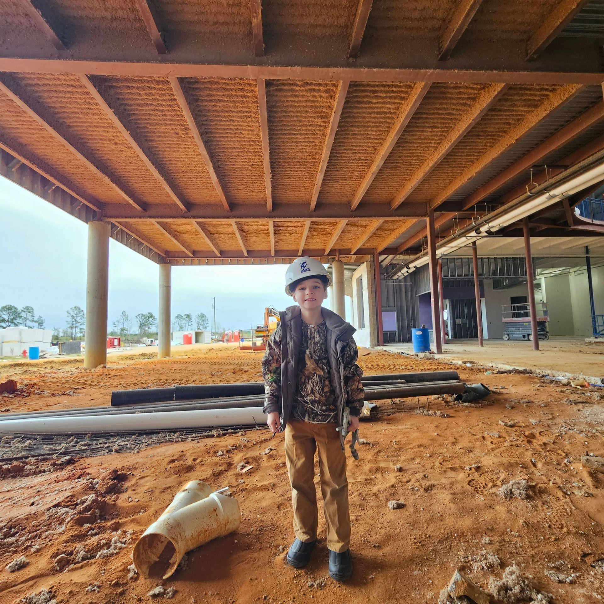 A person wearing a white hard hat stands on dirt ground under the exposed wooden ceiling of a construction site.