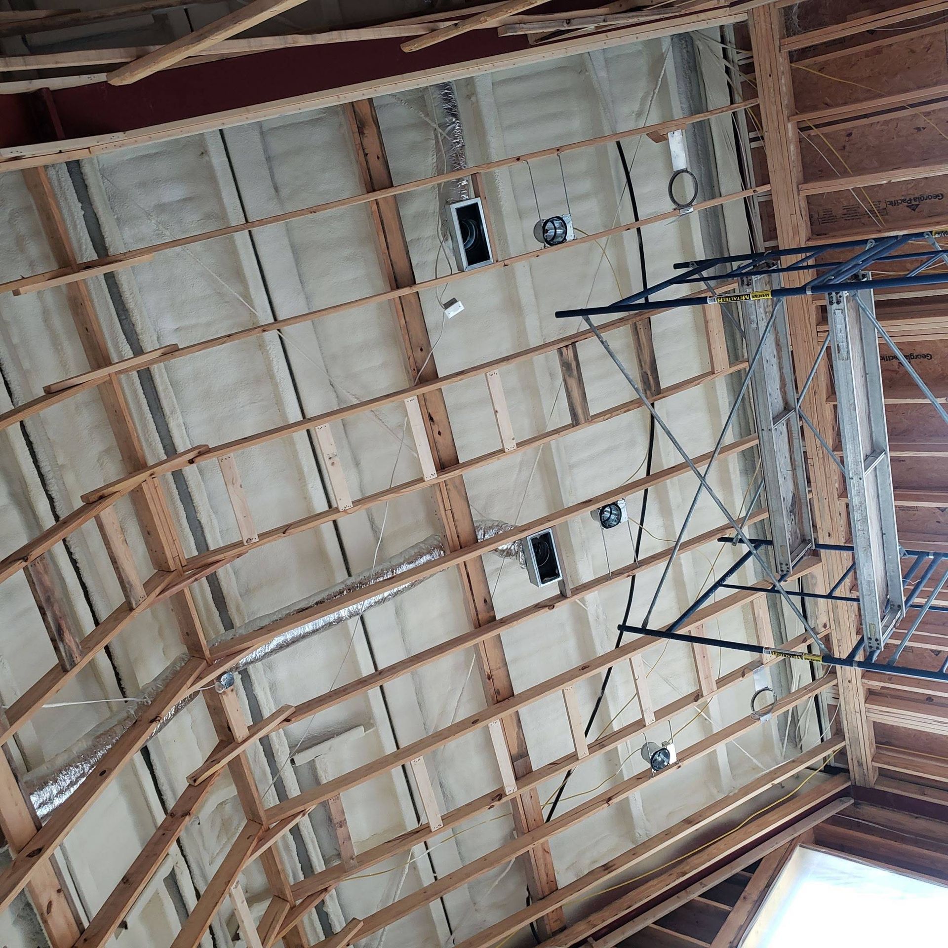 An unfinished room showing exposed wooden ceiling rafters, spray-foam insulation, electrical wiring, and metal scaffolding.