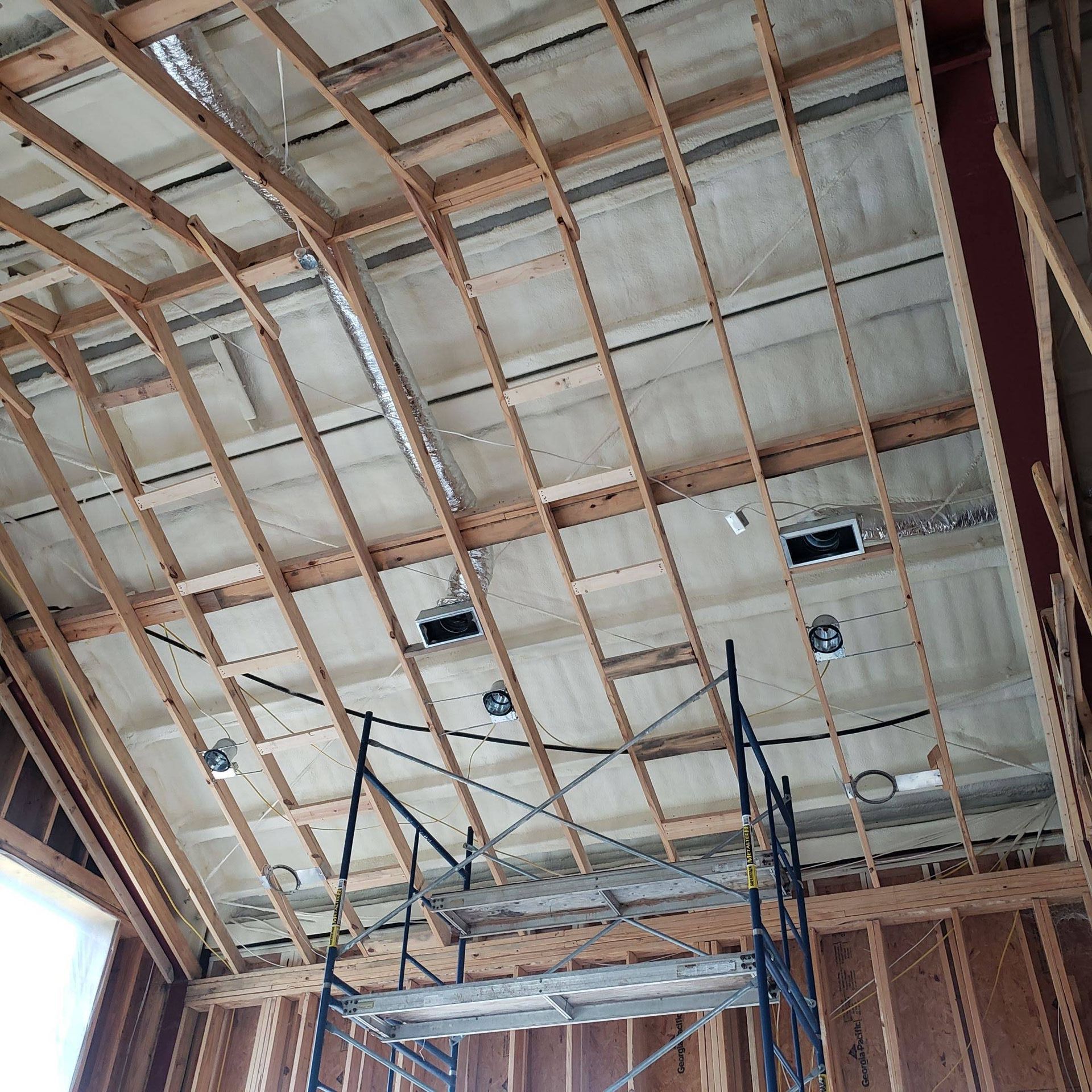 A construction site interior showing a sloped ceiling with exposed wood framing, light fixtures, and a rolling metal scaffold.