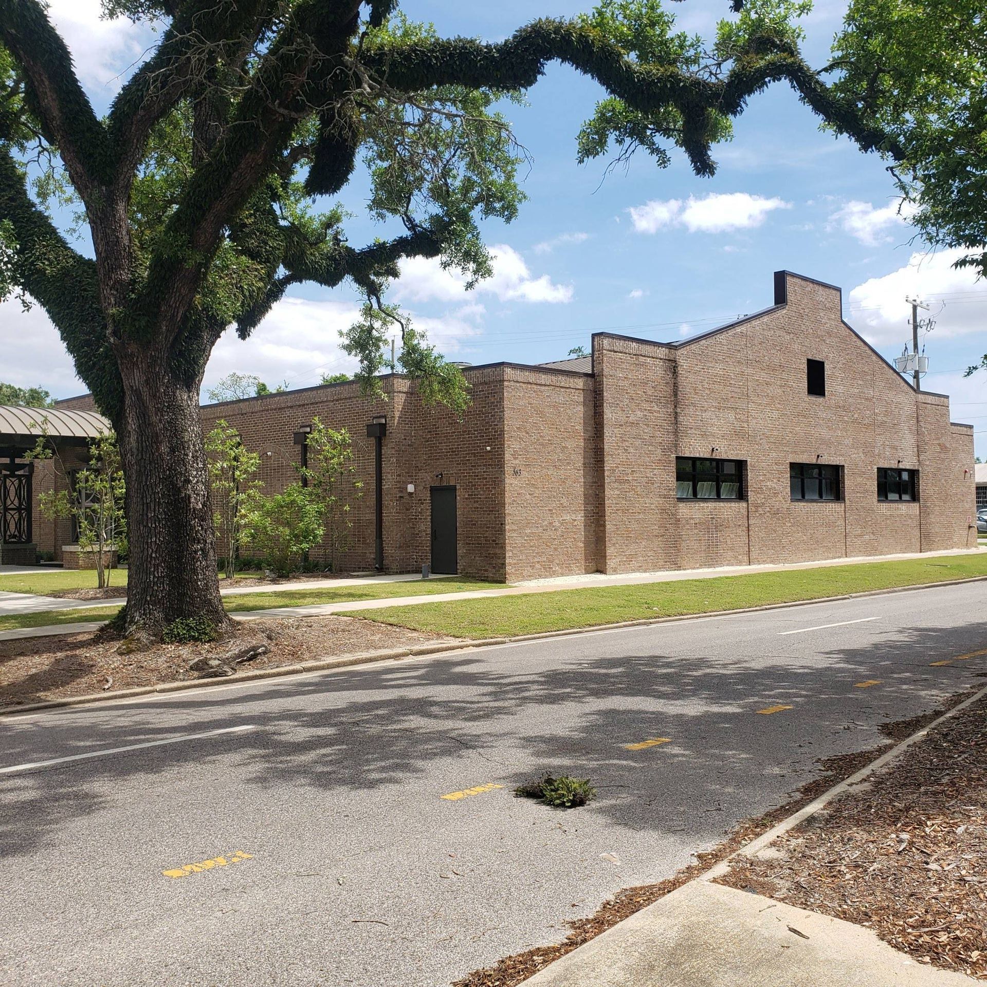 A low-rise, brown brick building on a street corner, partially shaded by a large tree under a sunny blue sky.