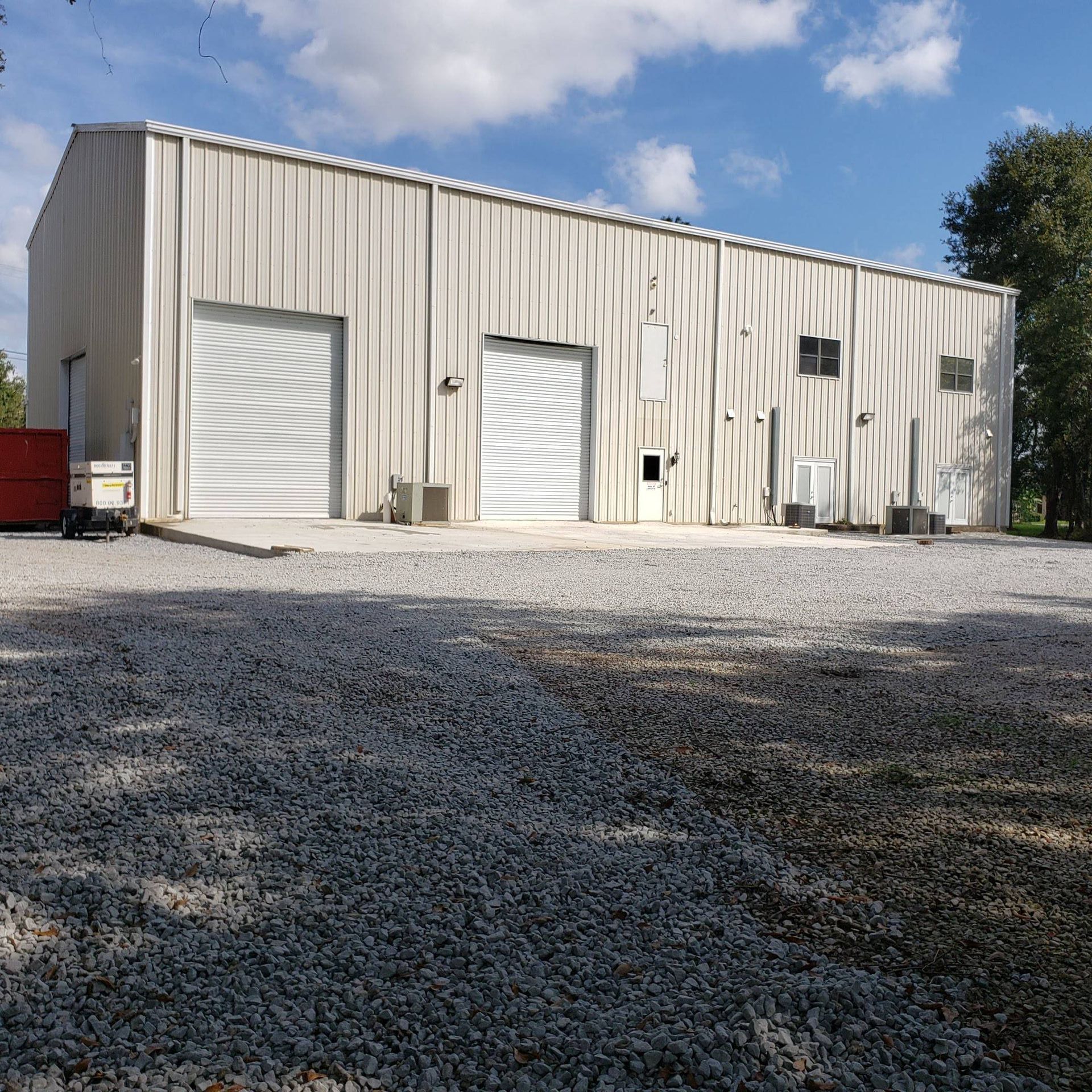 A large, light-colored metal industrial warehouse with two roll-up doors and a gravel lot under a blue, cloudy sky.