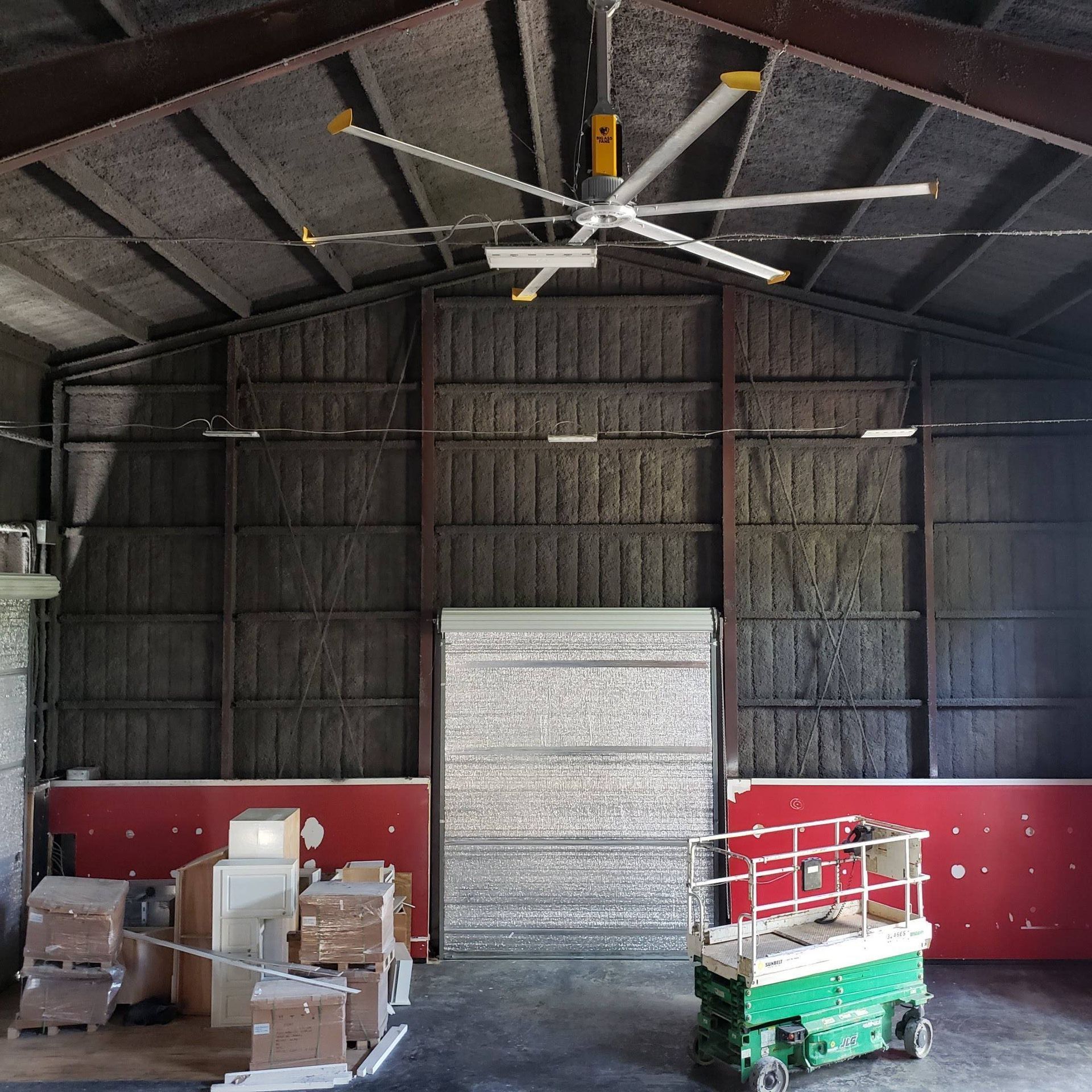 Interior view of a warehouse with a large overhead fan, stacked boxes on the left, and a scissor lift on the right.