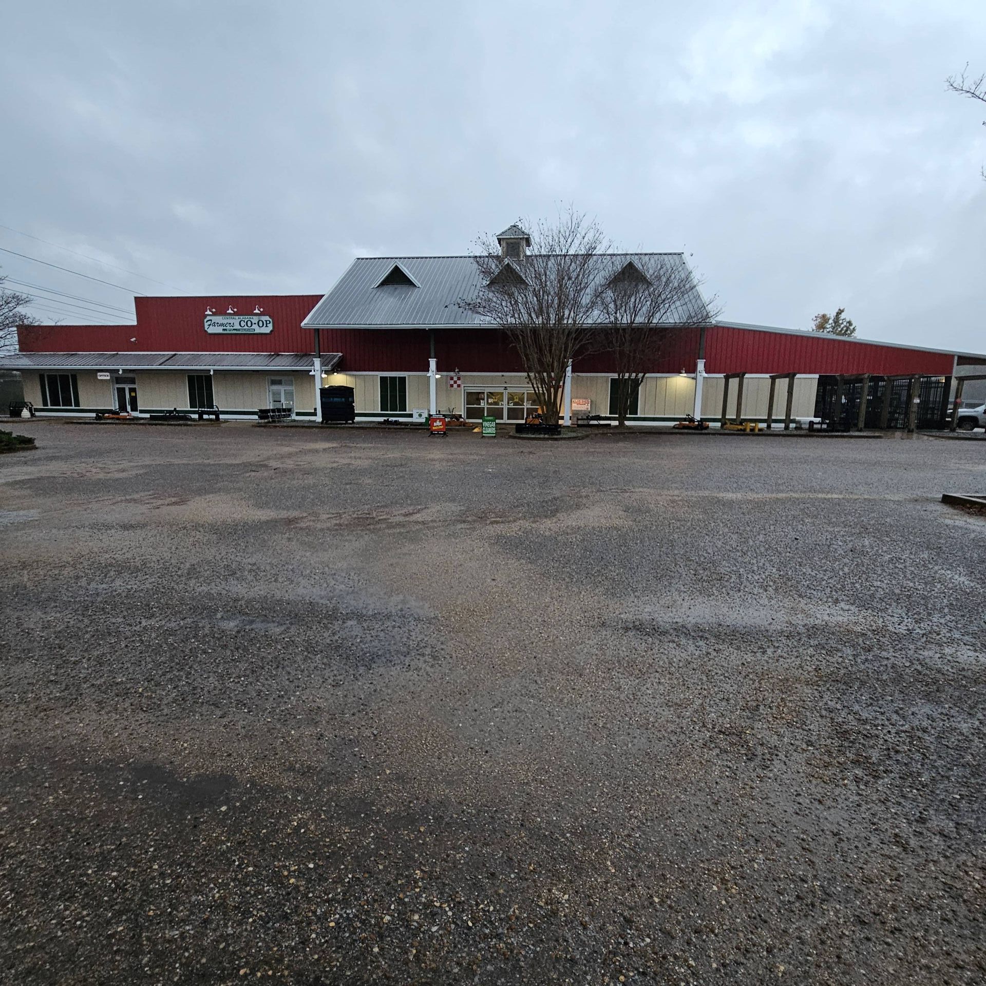 A wide, single-story building with a red exterior, metal roof, and large gravel parking lot under a cloudy sky.