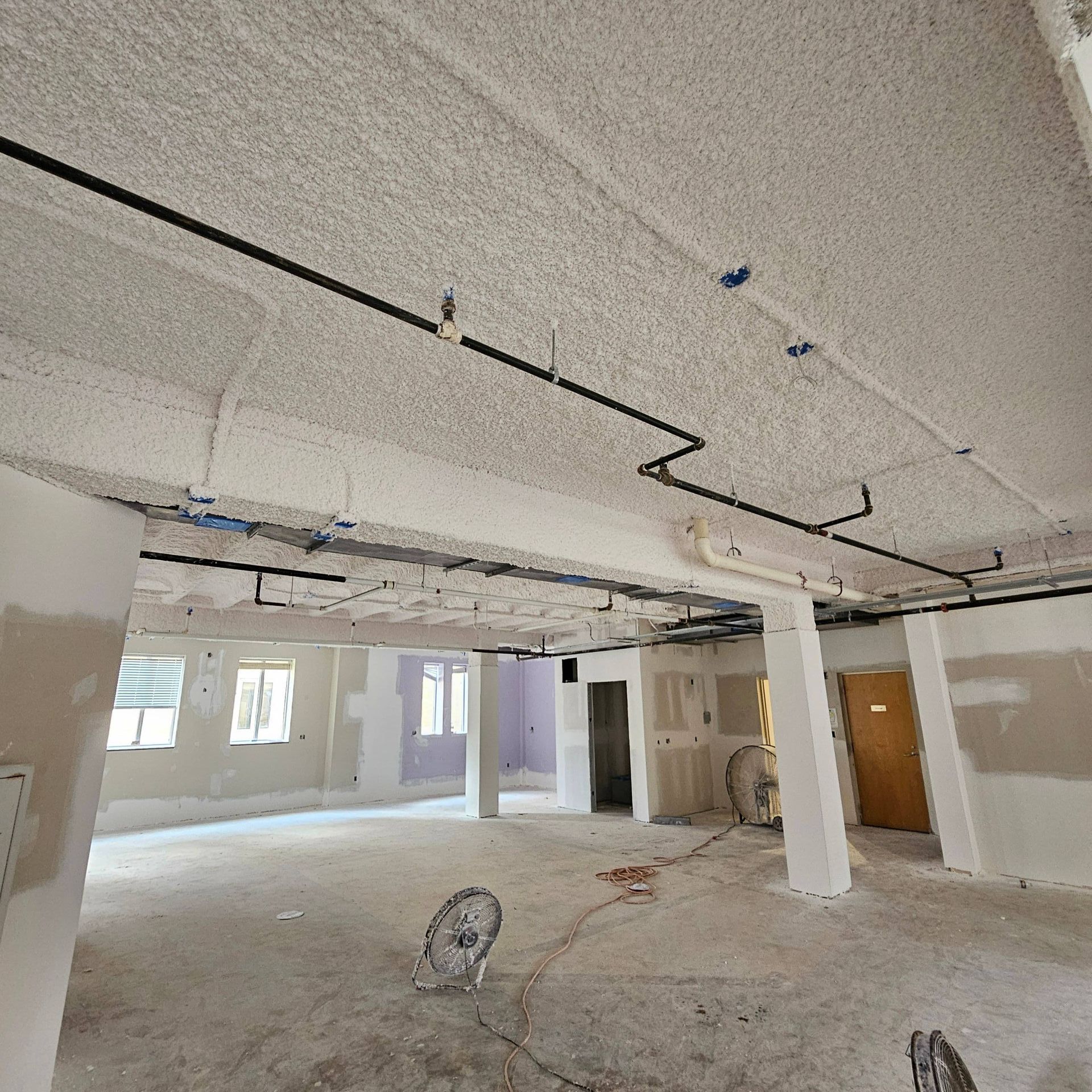 An unfinished commercial interior space with exposed ceilings, black piping, white columns, and construction materials.
