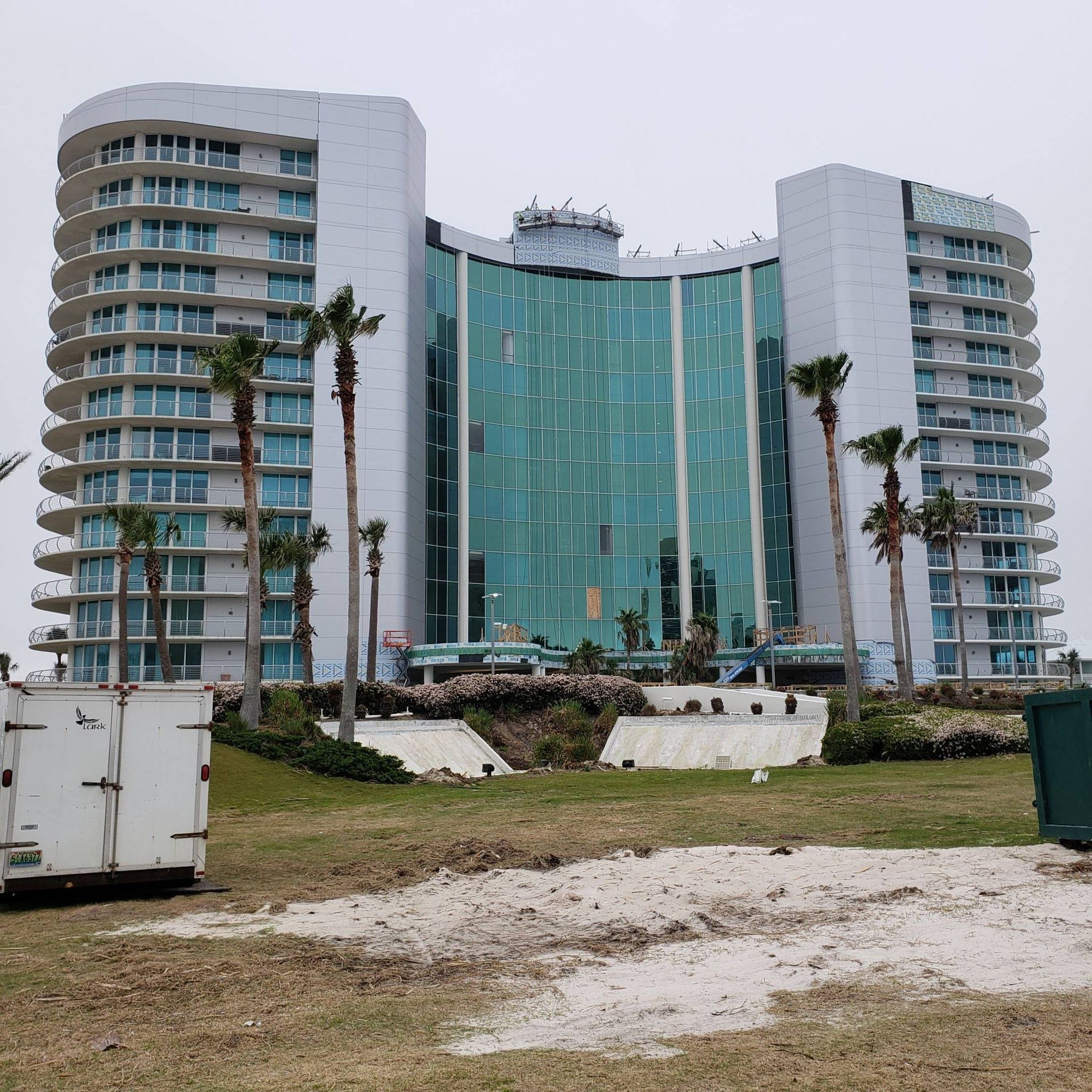 A large, multi-story white hotel with glass walls, surrounded by palm trees under a cloudy sky.
