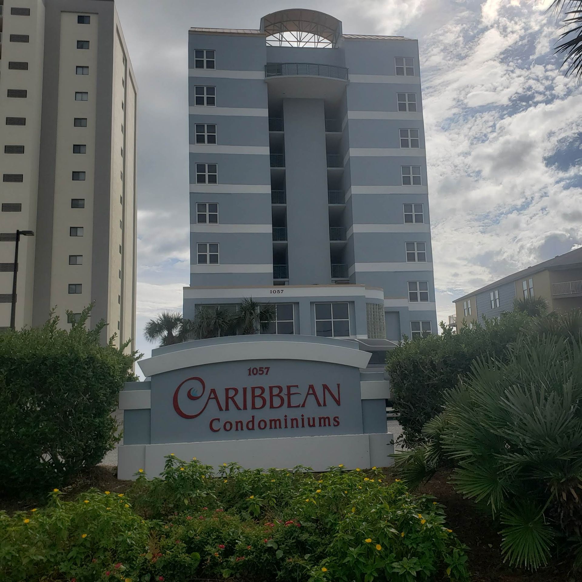 A light blue, multi-story Caribbean Condominium building with a prominent sign in the foreground amidst greenery.