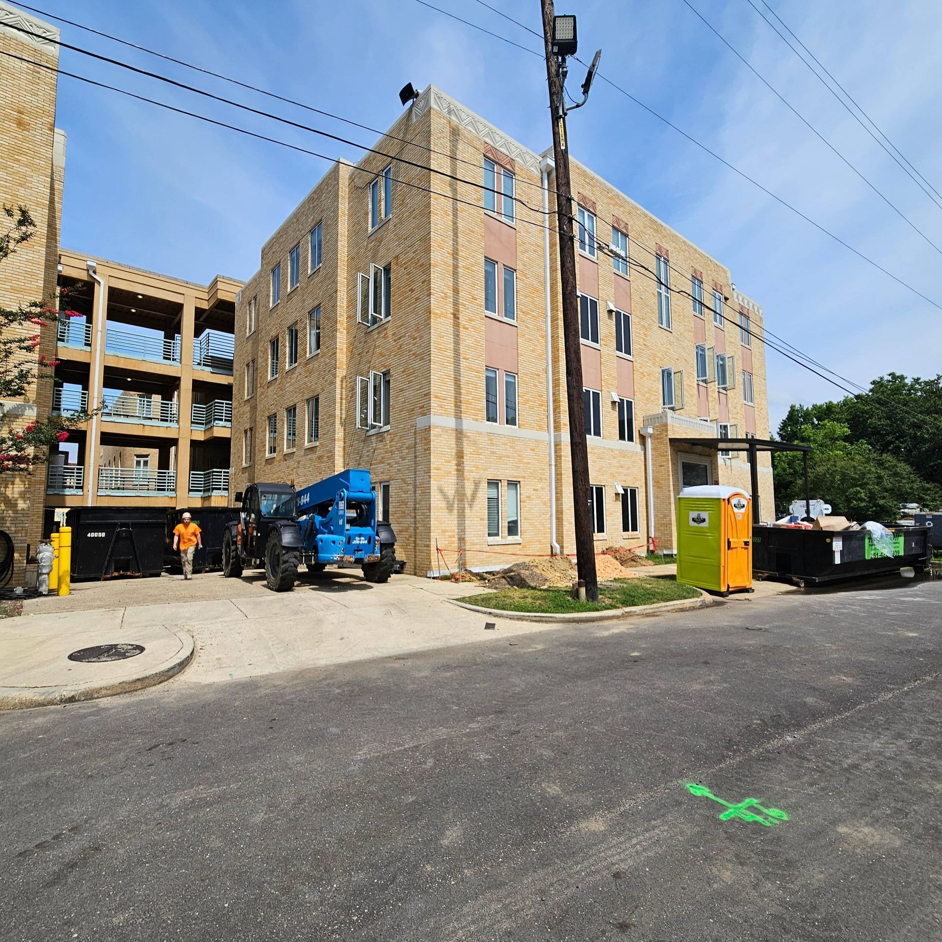 A construction site featuring a tan brick apartment building, heavy machinery, a portable toilet, and a dump truck.