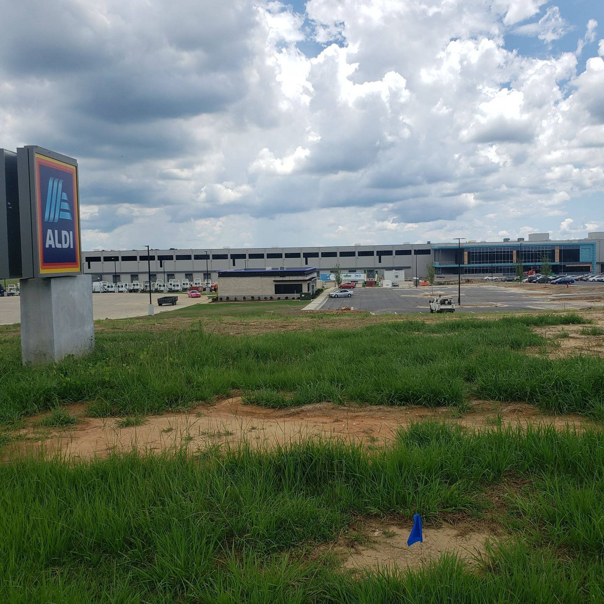 An Aldi sign stands in the foreground of a grassy field, with a commercial plaza and a cloudy blue sky in the distance.