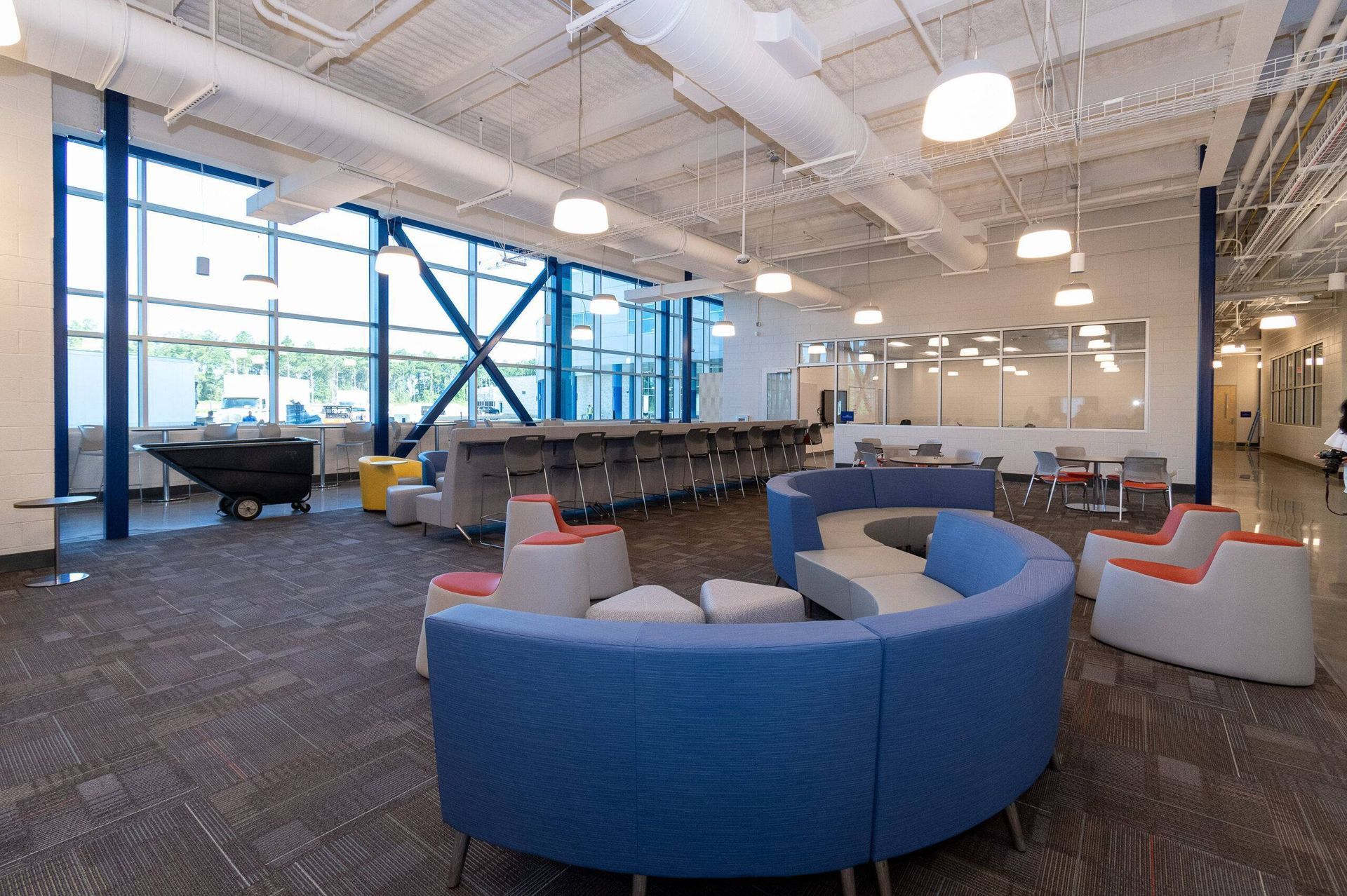 Modern office lounge with curved blue sofas, gray chairs, large windows, and an industrial-style ceiling.