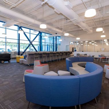 Modern office lounge with curved blue seating, stools, and bar-style tables in front of large windows with metal bracing.