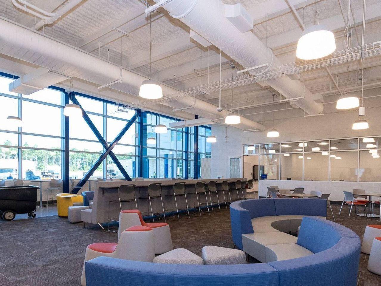 A modern office lounge featuring blue curved sectional seating, stools with orange tops, and a long window-facing desk.