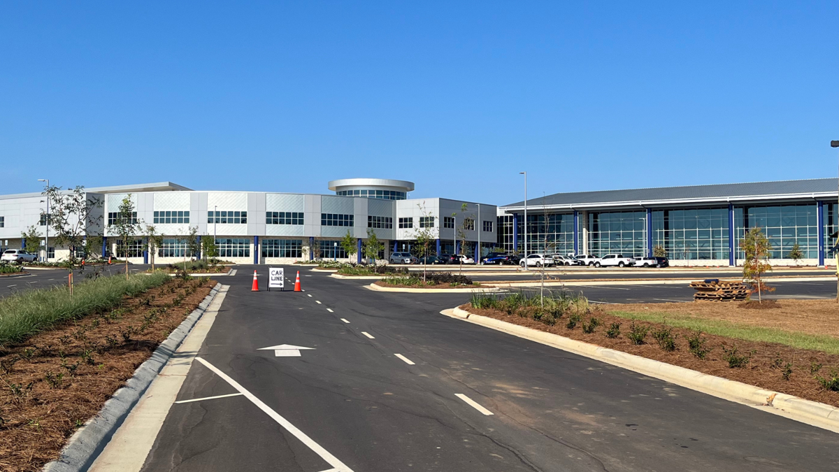 A paved road leads toward a modern, light-colored commercial building with large windows under a clear blue sky.