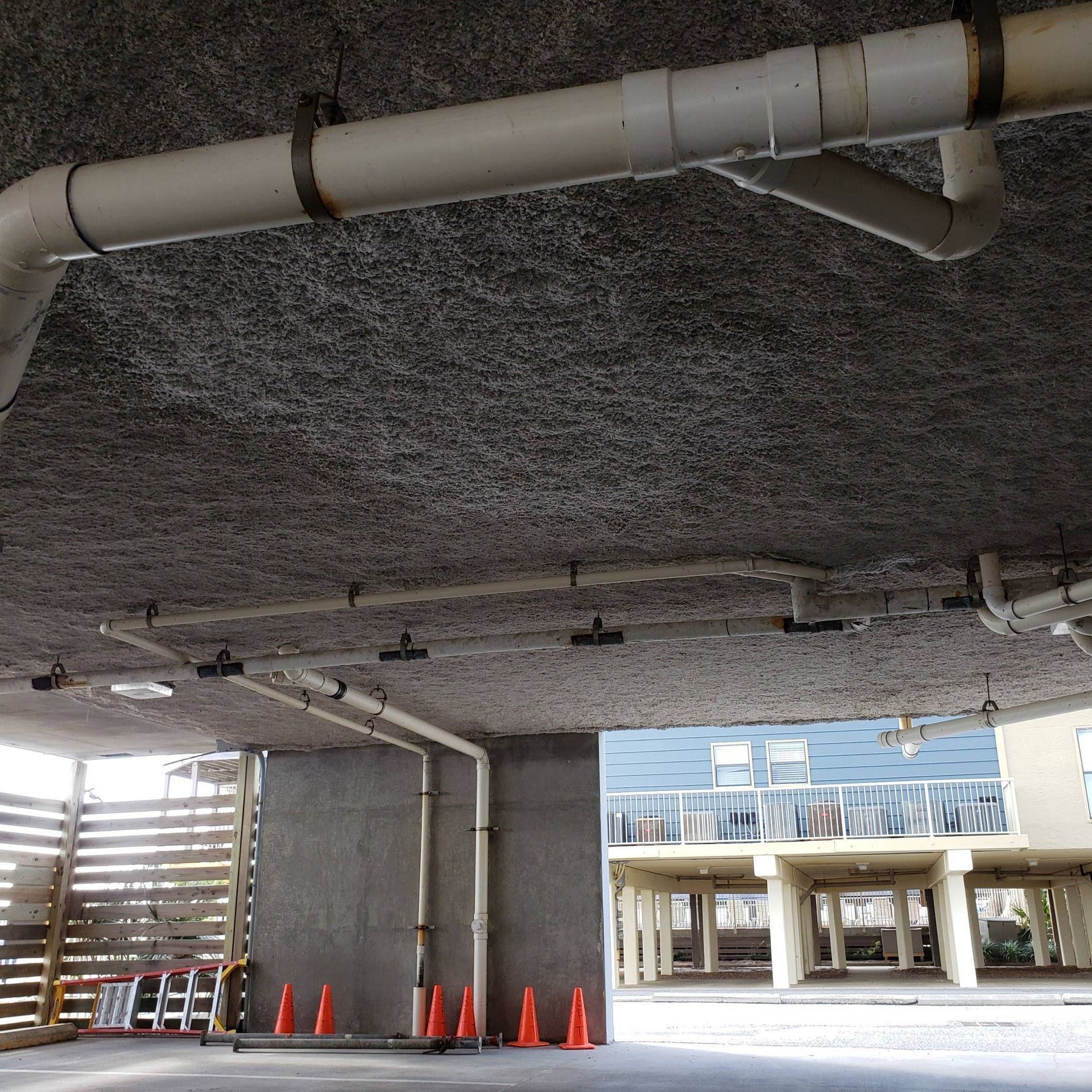 An outdoor parking area under a building with exposed pipes on the textured ceiling and several orange traffic cones.