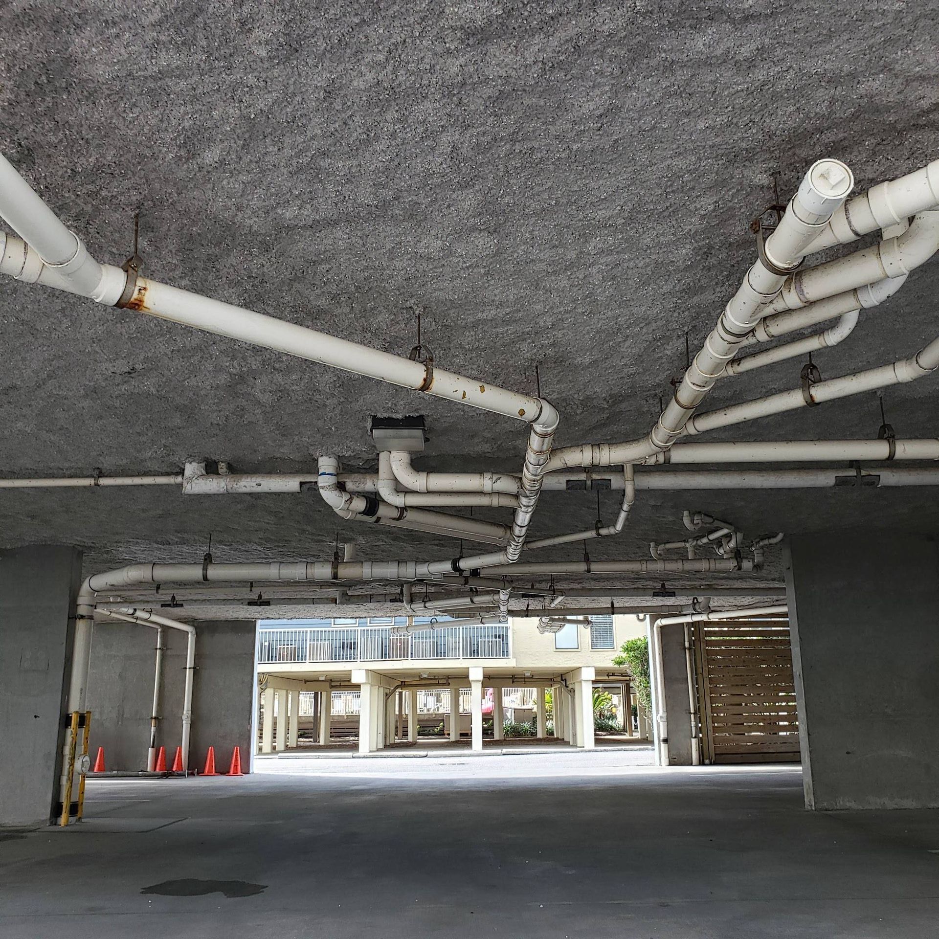 A low-angle view of a concrete ceiling in a parking structure with a complex network of white PVC pipes mounted to it.