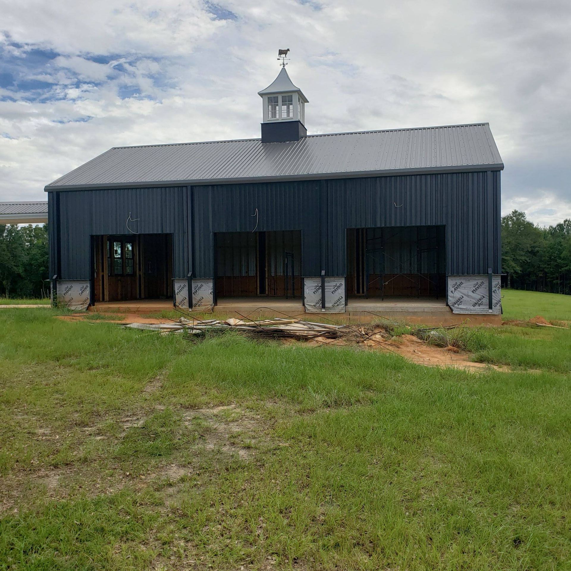 A new, dark gray metal barn with three open bays and a small cupola sits on a grassy field under a cloudy sky.
