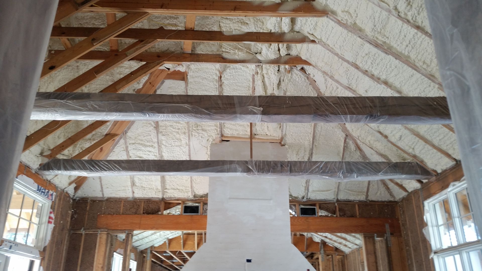 An interior view of a room under construction, featuring exposed wooden beams, a central fireplace, and spray-foam walls.
