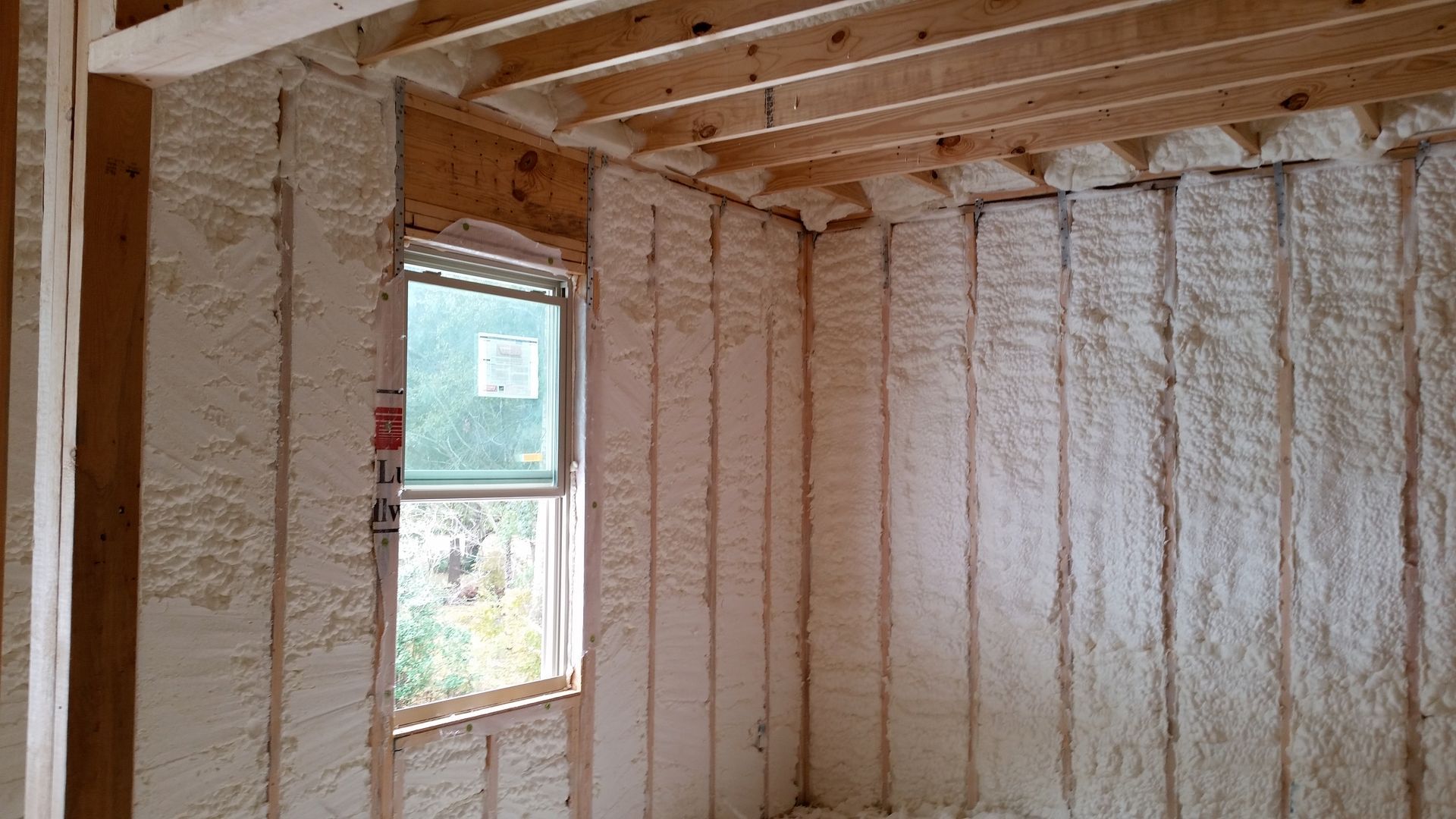 Interior room under construction with white spray foam insulation applied between wooden wall studs and ceiling joists.