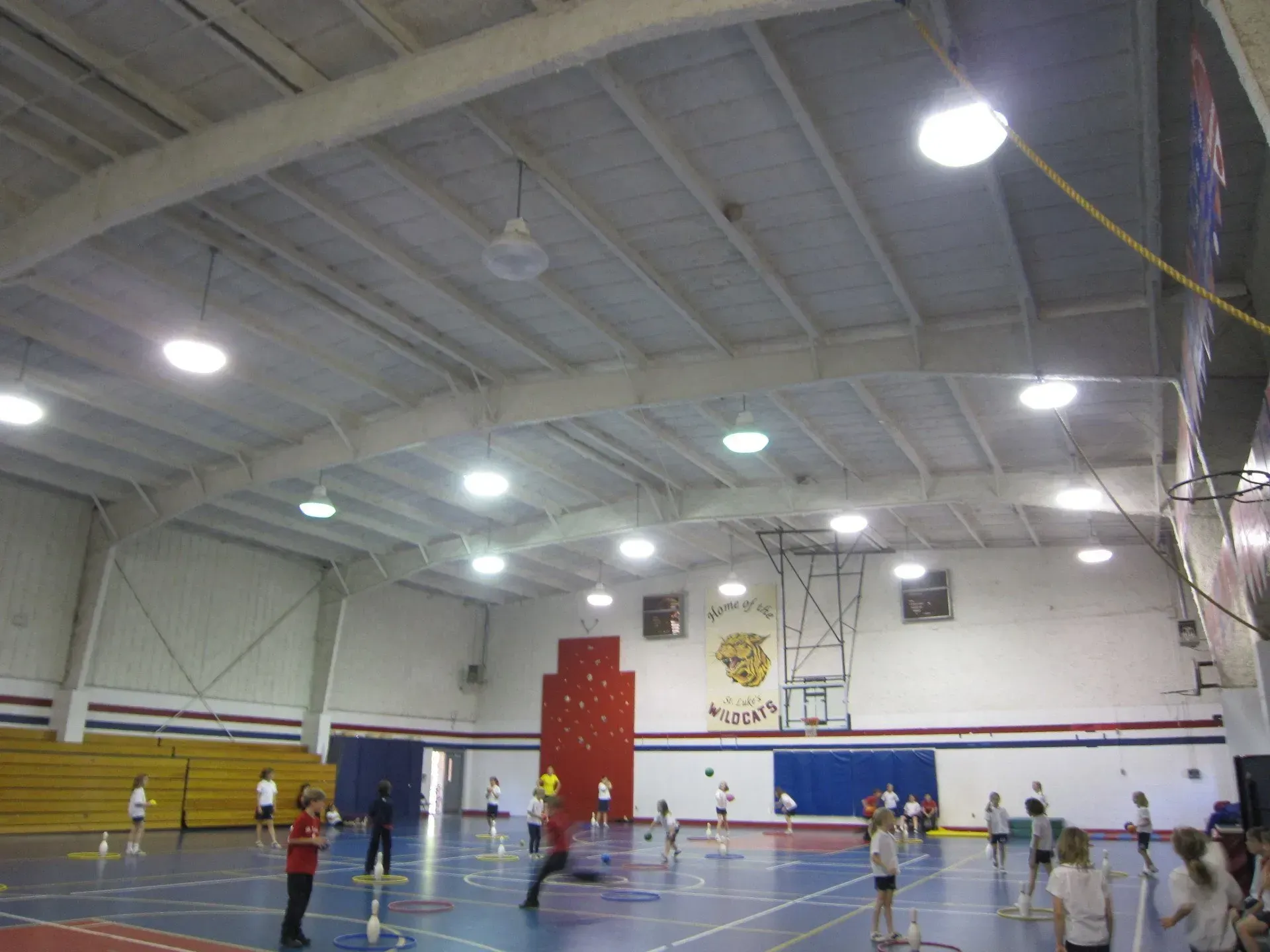 Children play on a blue indoor basketball court with red and blue wall stripes and a ceiling with multiple light fixtures.