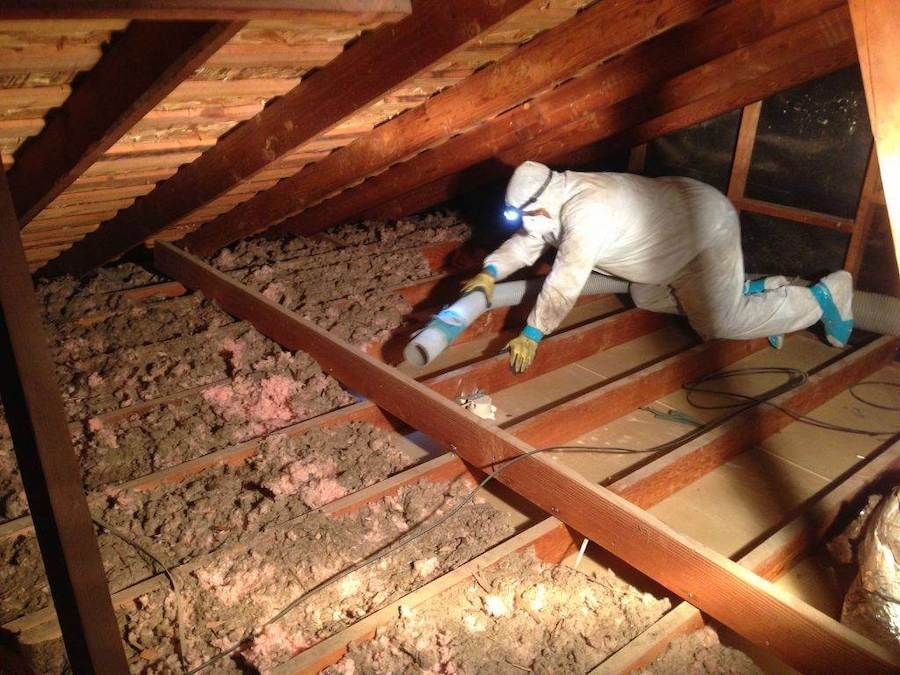 A worker in protective gear and a headlamp blows loose-fill insulation between wooden rafters in an attic.