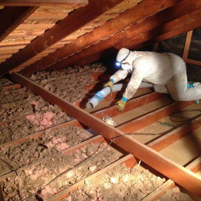A person in protective coveralls uses a hose to blow insulation into a wooden attic space.