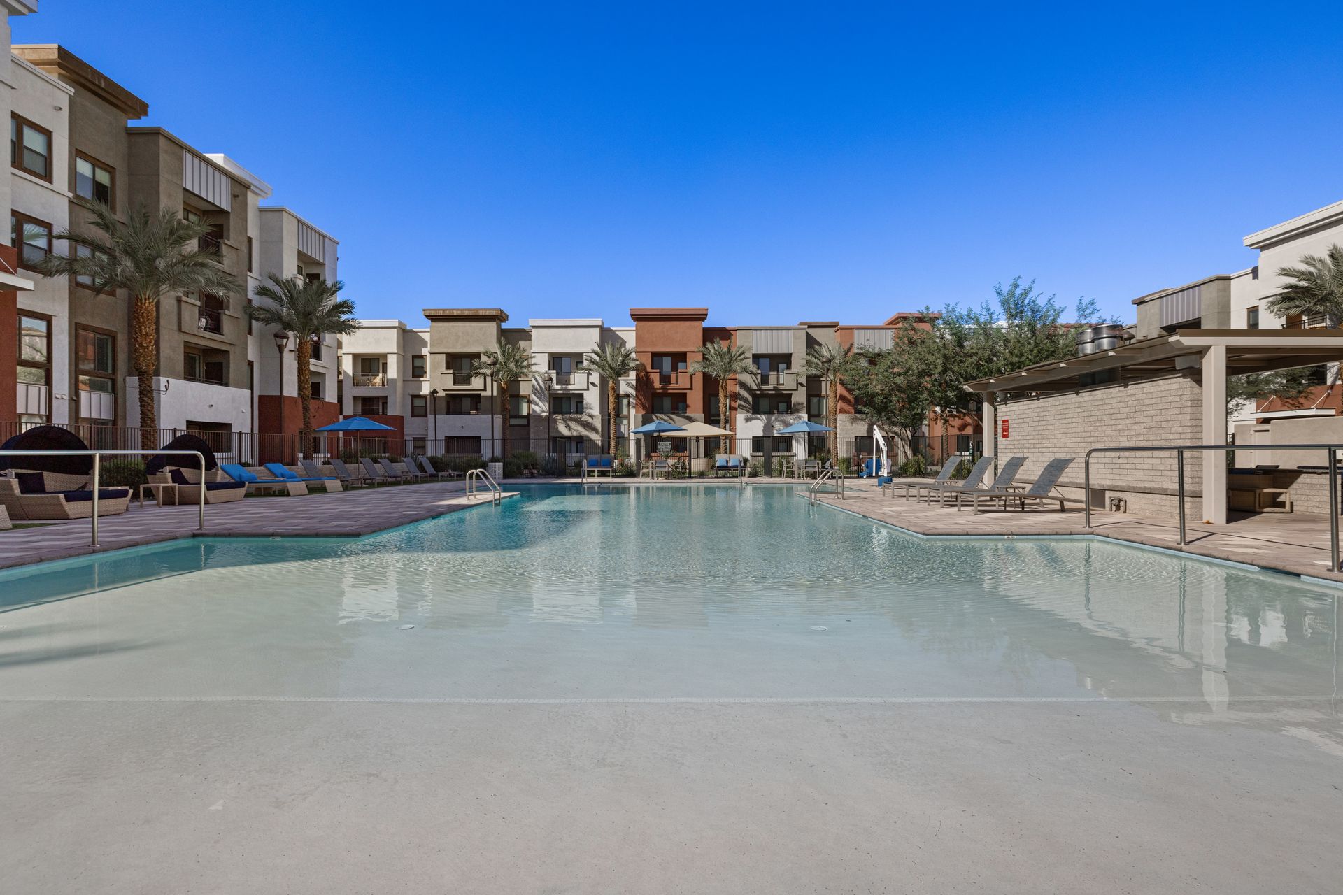 Modern apartment courtyard with a large swimming pool, lounge chairs, and clear blue sky