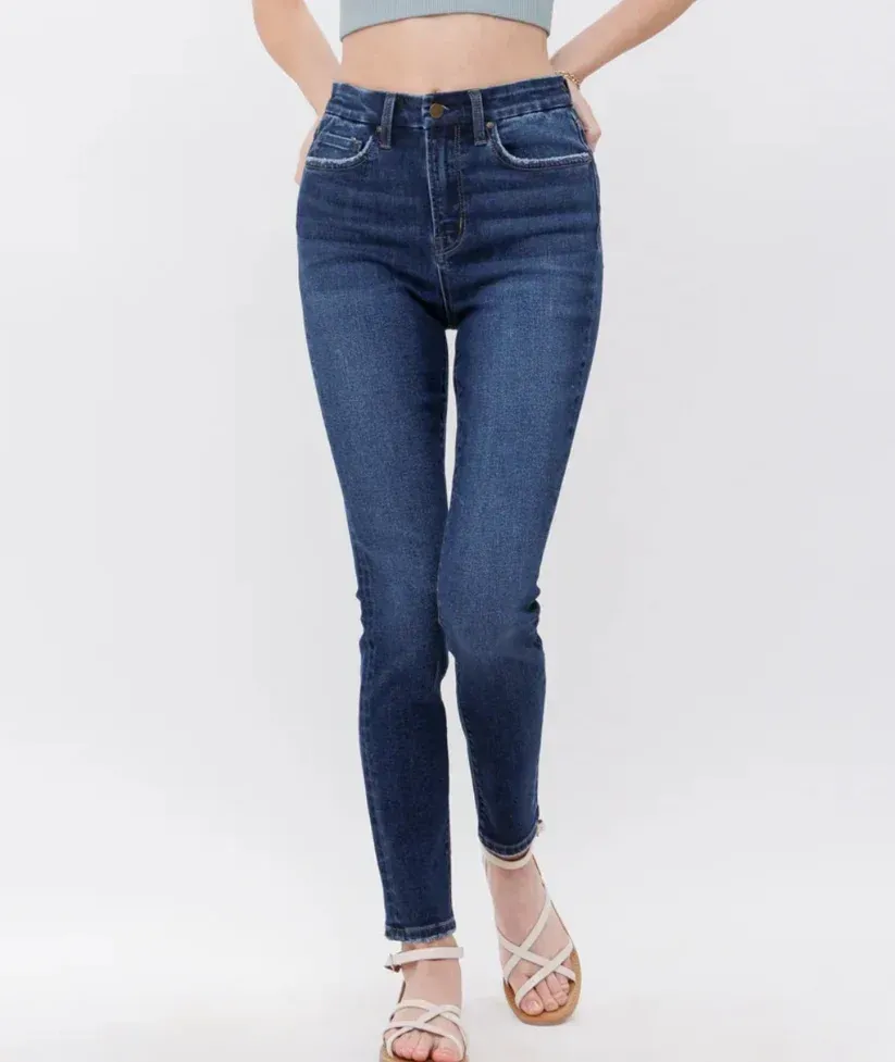Model wearing dark blue skinny jeans and white heeled sandals, standing against a light background