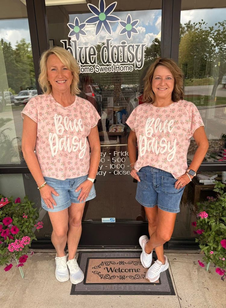 Woman standing outside a storefront, smiling, in a maroon polka-dot top, jeans, and sandals.