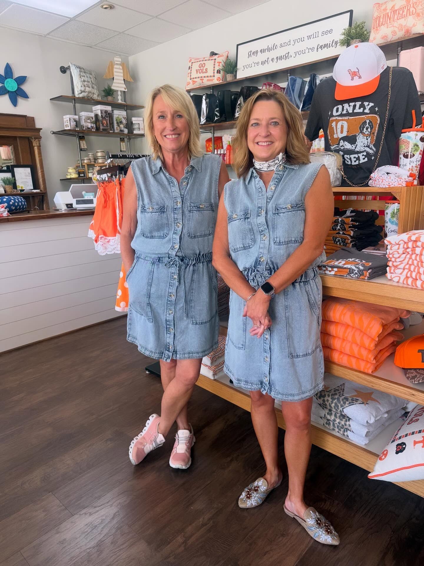 Two women in matching pink tops and denim shorts stand smiling outside a shop entrance.