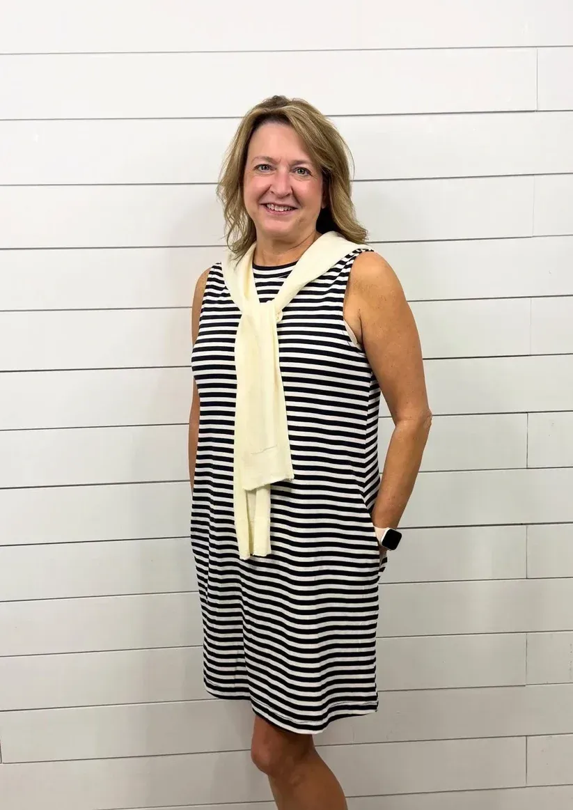 Woman in a black-and-white striped dress and cream scarf standing against a white brick wall.