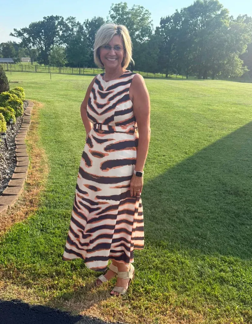 Woman in a striped dress standing on a grassy lawn beside a stone path, smiling in sunlight.