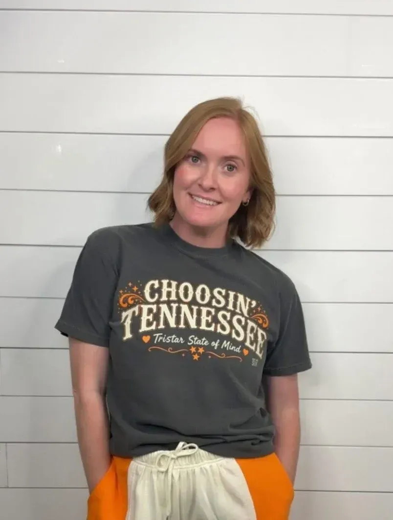 Person standing against a white wall, wearing a gray “Choosing Tennessee” T-shirt and orange shorts.