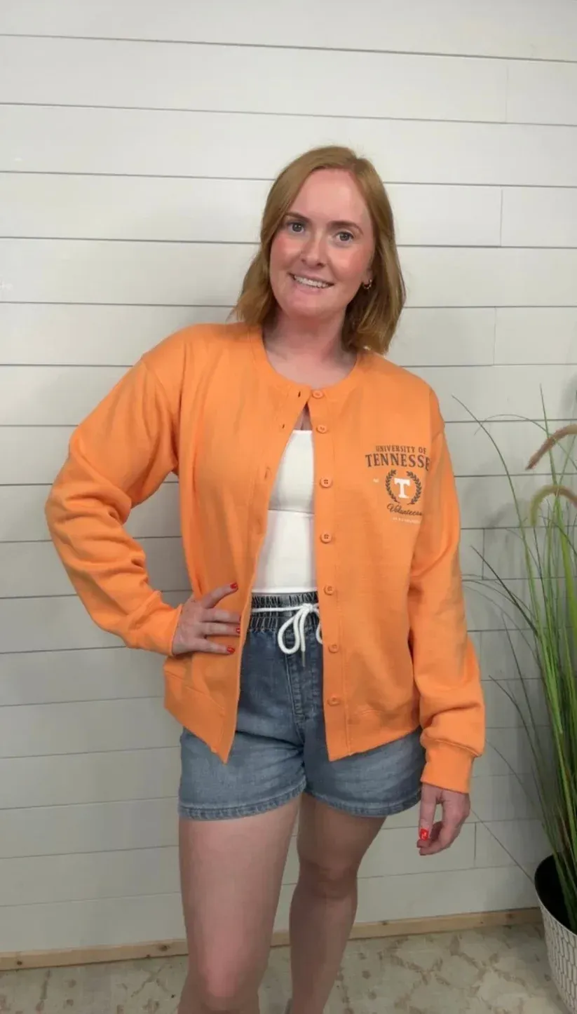 Woman standing indoors against a white wall, wearing an orange jacket, denim shorts, and smiling.