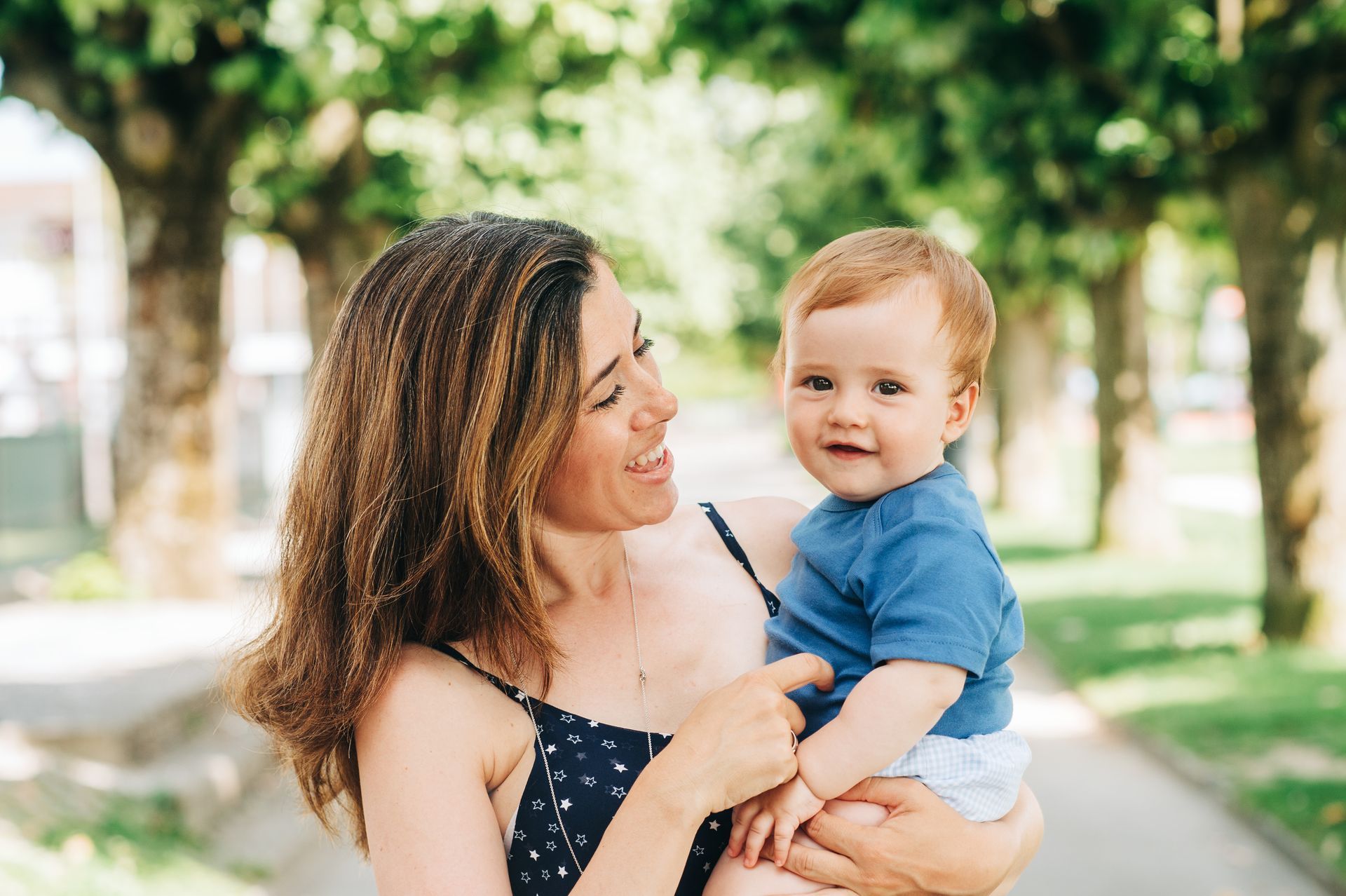 Woman holding a baby outside, smiling. Baby smiles, wearing blue shirt, looking at camera. Trees in background.