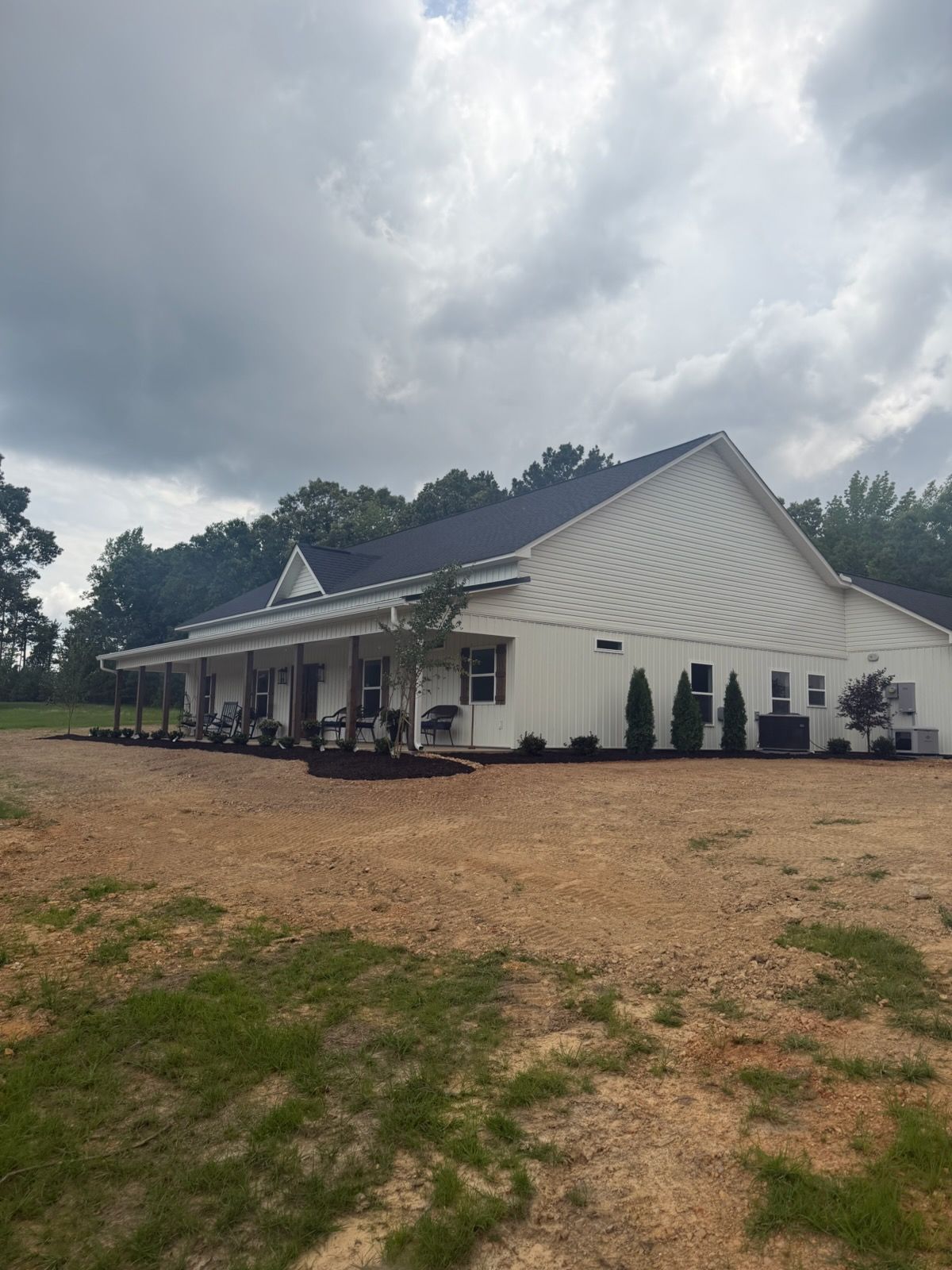 White building with dark roof on a gravel lot under cloudy skies, bordered by trees.