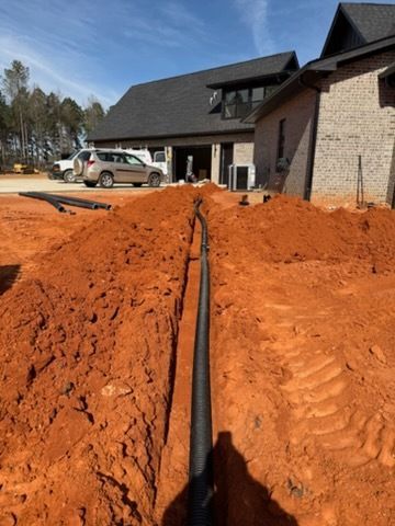 Trench with black pipe laid in red dirt beside a house under construction on a sunny day