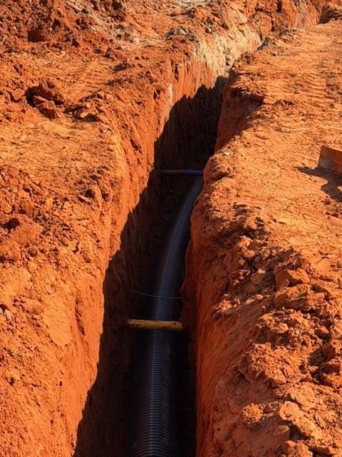 Narrow irrigation trench cut through red soil, with water flowing beneath a wooden plank.