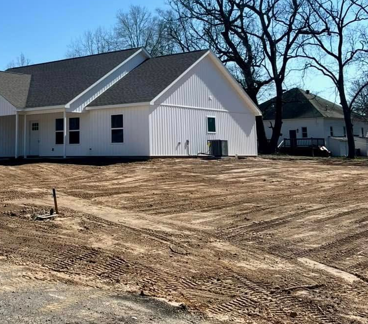 New white house under construction beside a dirt lot and leafless trees under a blue sky