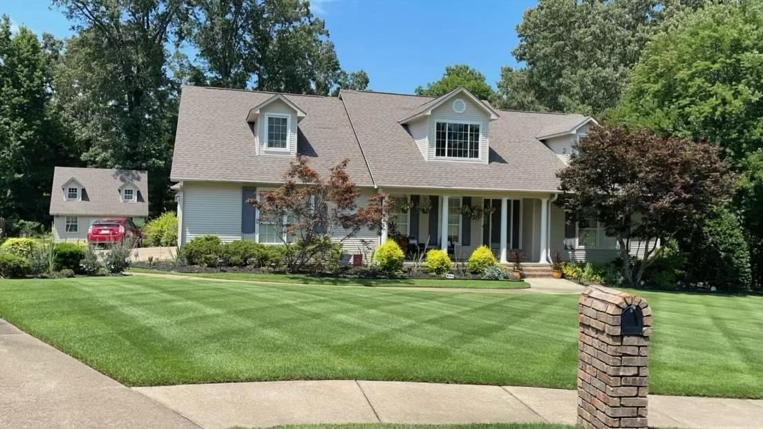 Suburban white house with gray roof, flower beds, and a manicured green lawn on a sunny day
