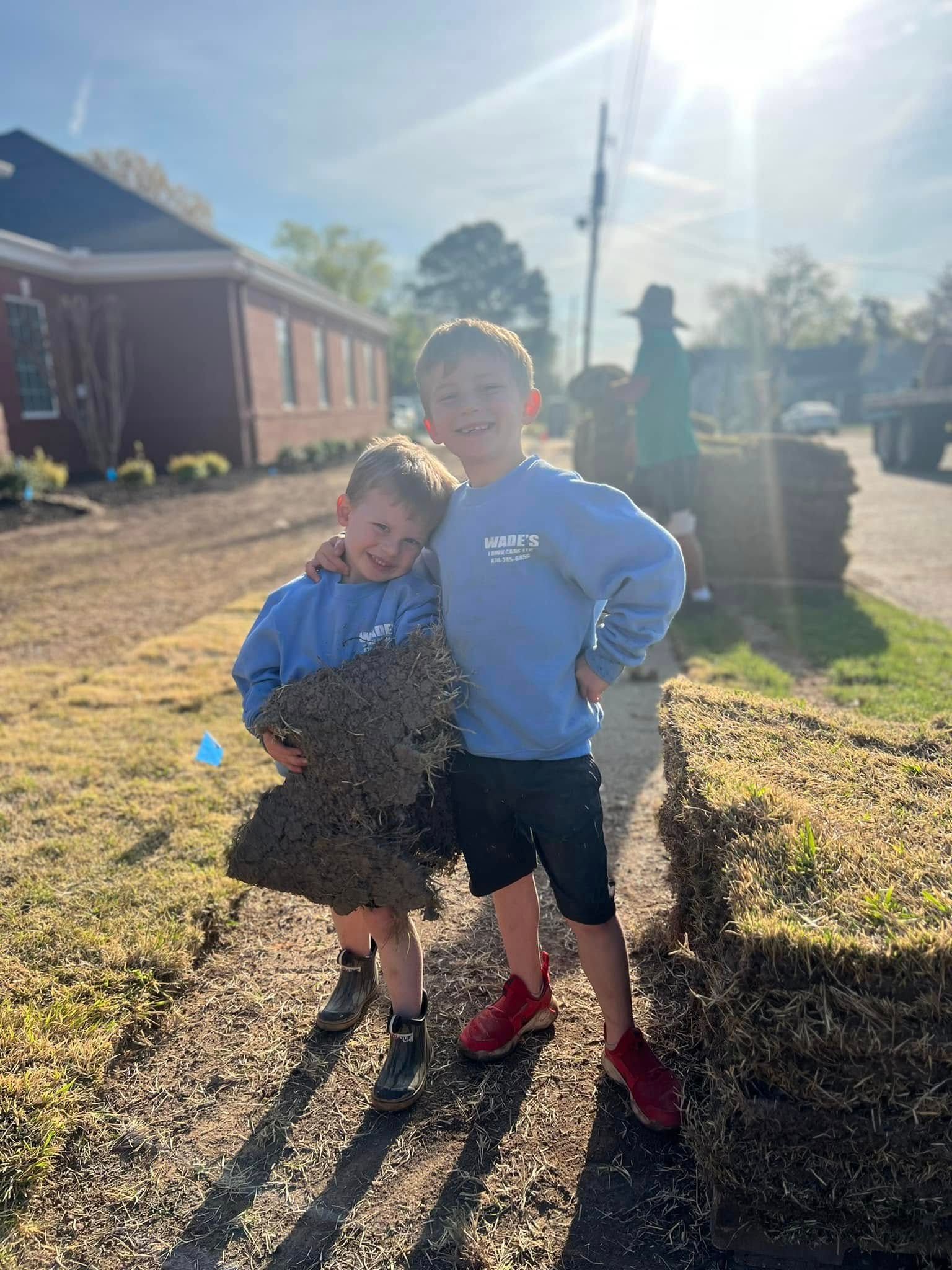 Two children standing together outdoors in bright sunlight beside a brick building and sidewalk.