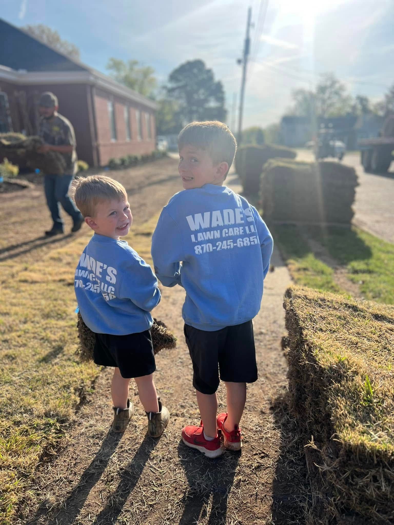 Two children in blue shirts stand on a sunny sidewalk near a brick building and a parked truck.