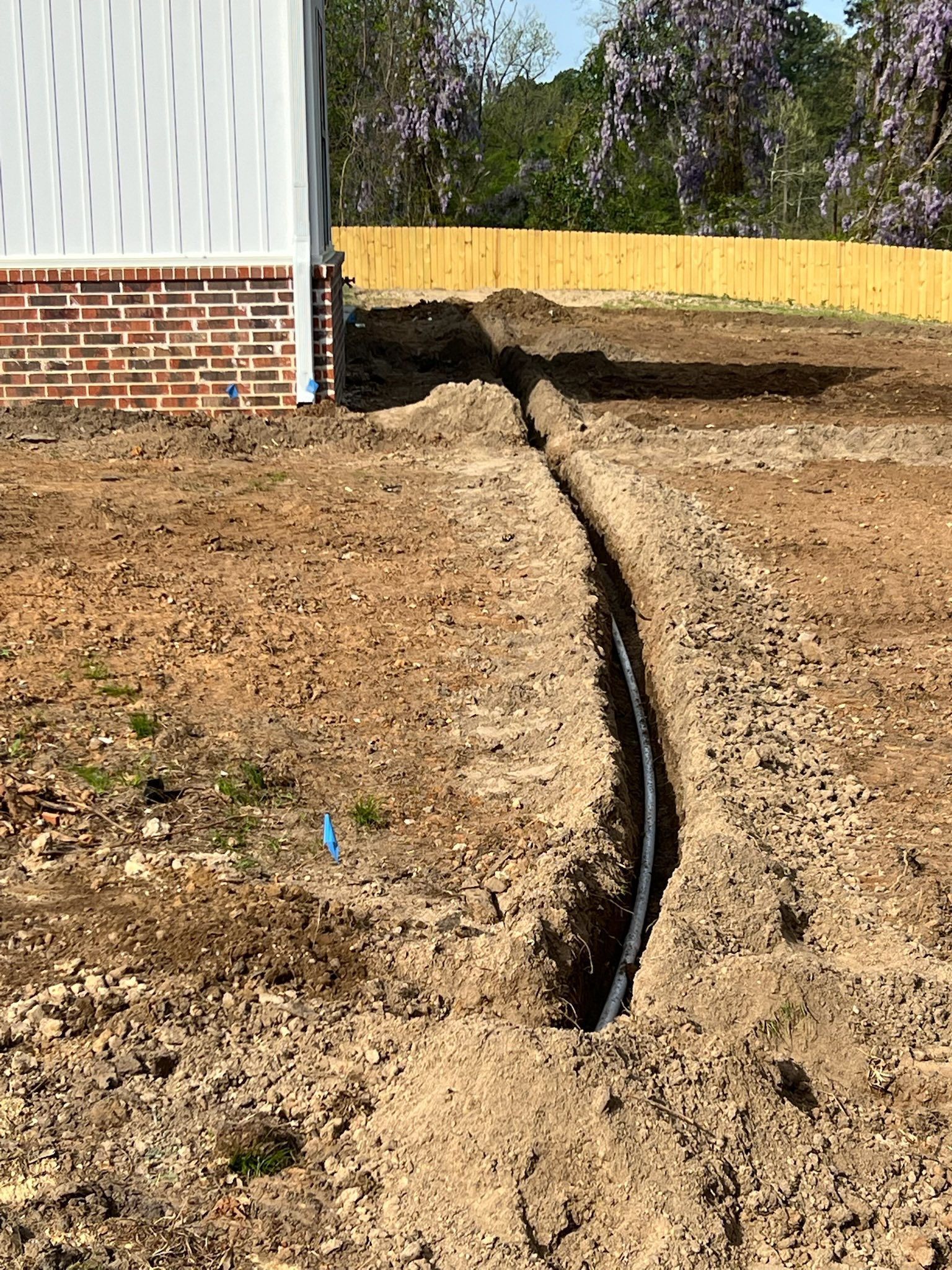 Wide trench cut through a dirt yard beside a brick house, leading toward a yellow fence.