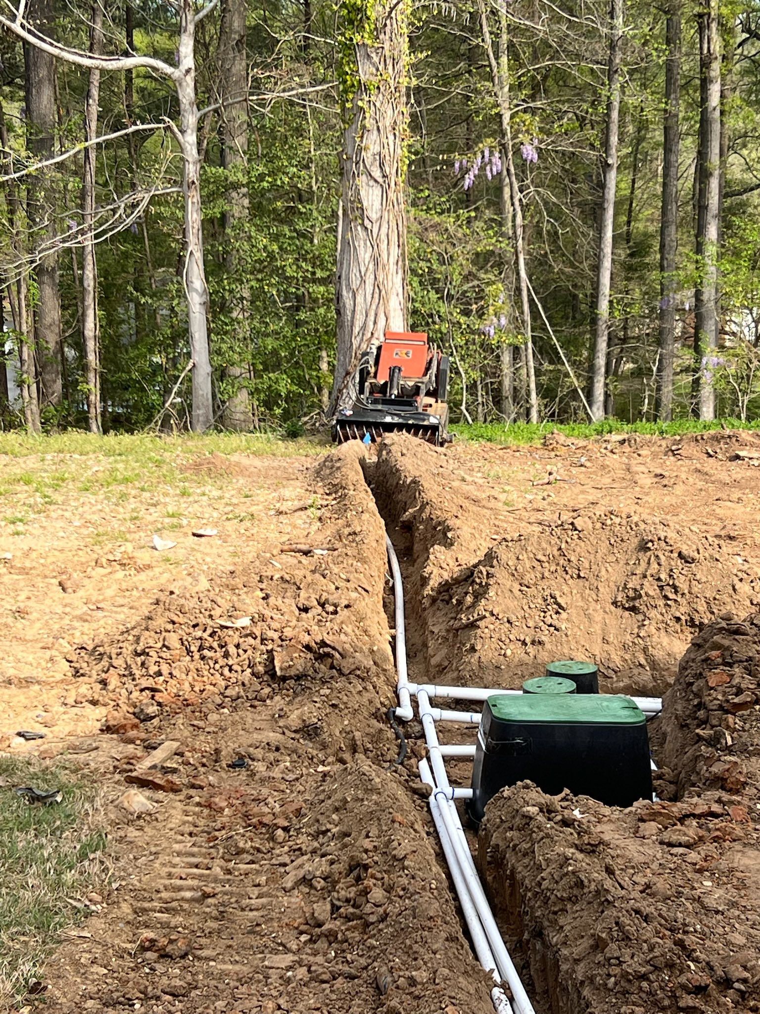 Orange trenching machine digging a long ditch in a wooded field beside a buried green pipe