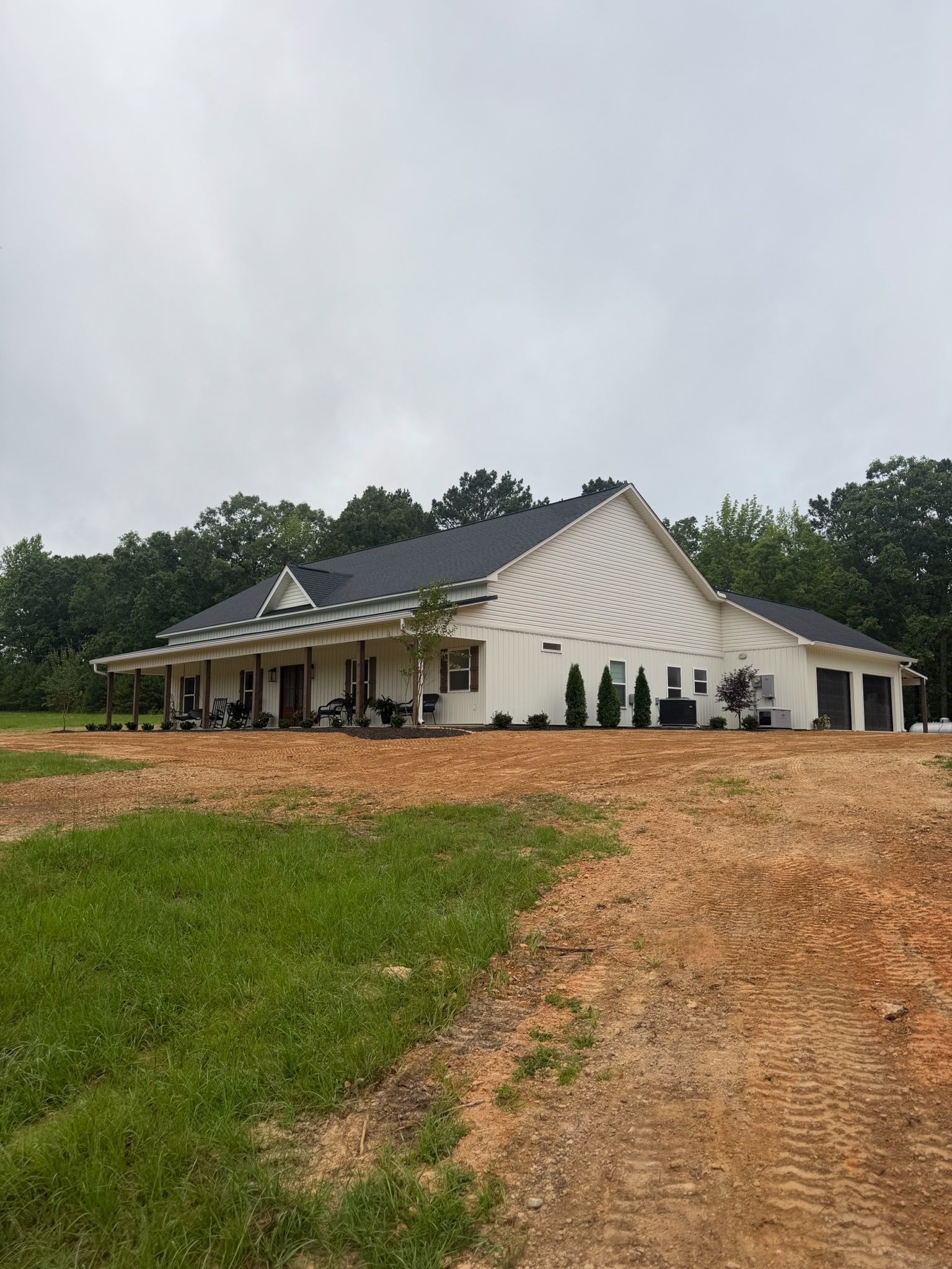 White house with gabled roof on a muddy lot under an overcast sky, surrounded by trees.