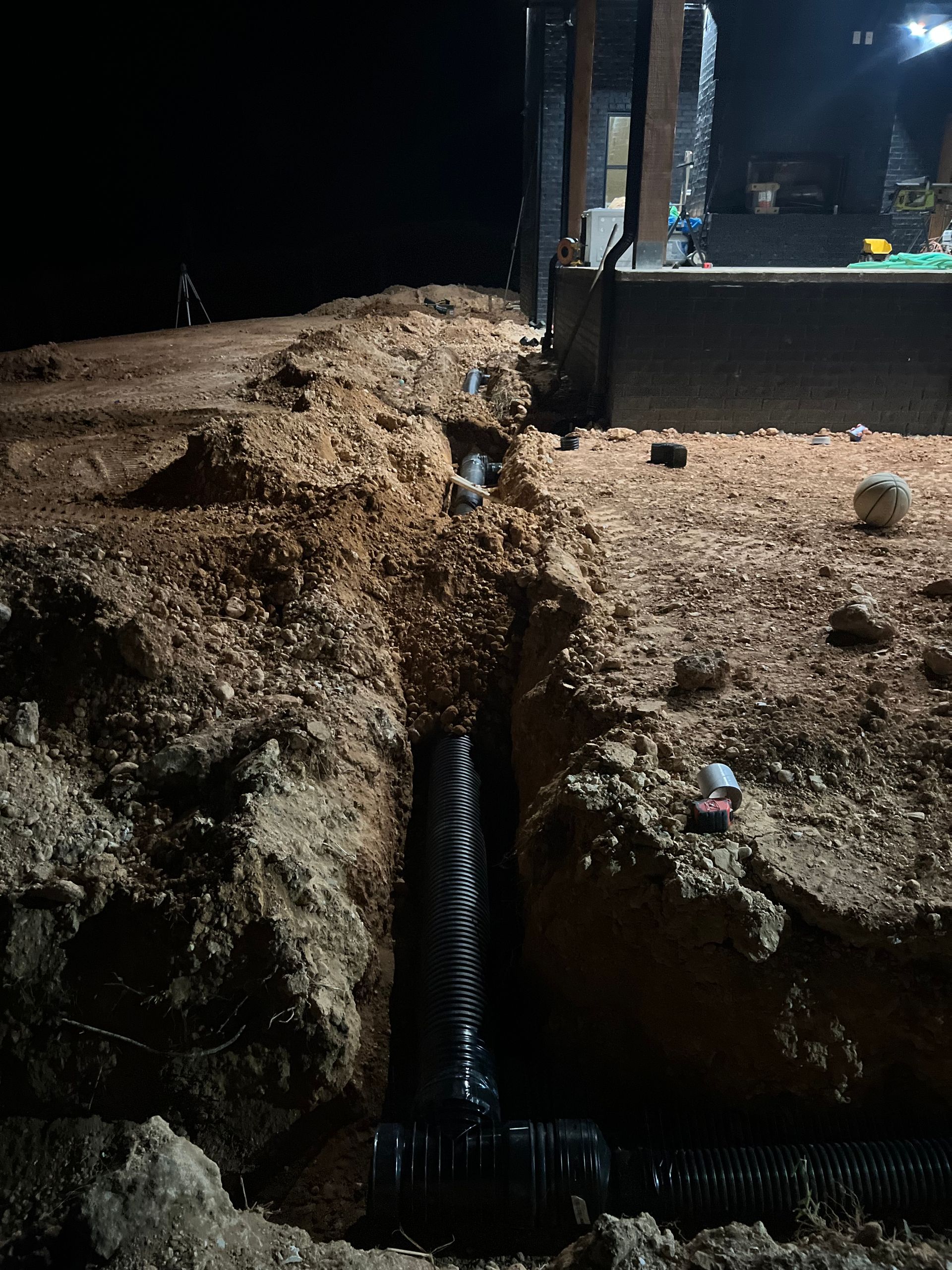 Muddy trench with a black corrugated pipe leading to a foundation opening beside a building exterior.