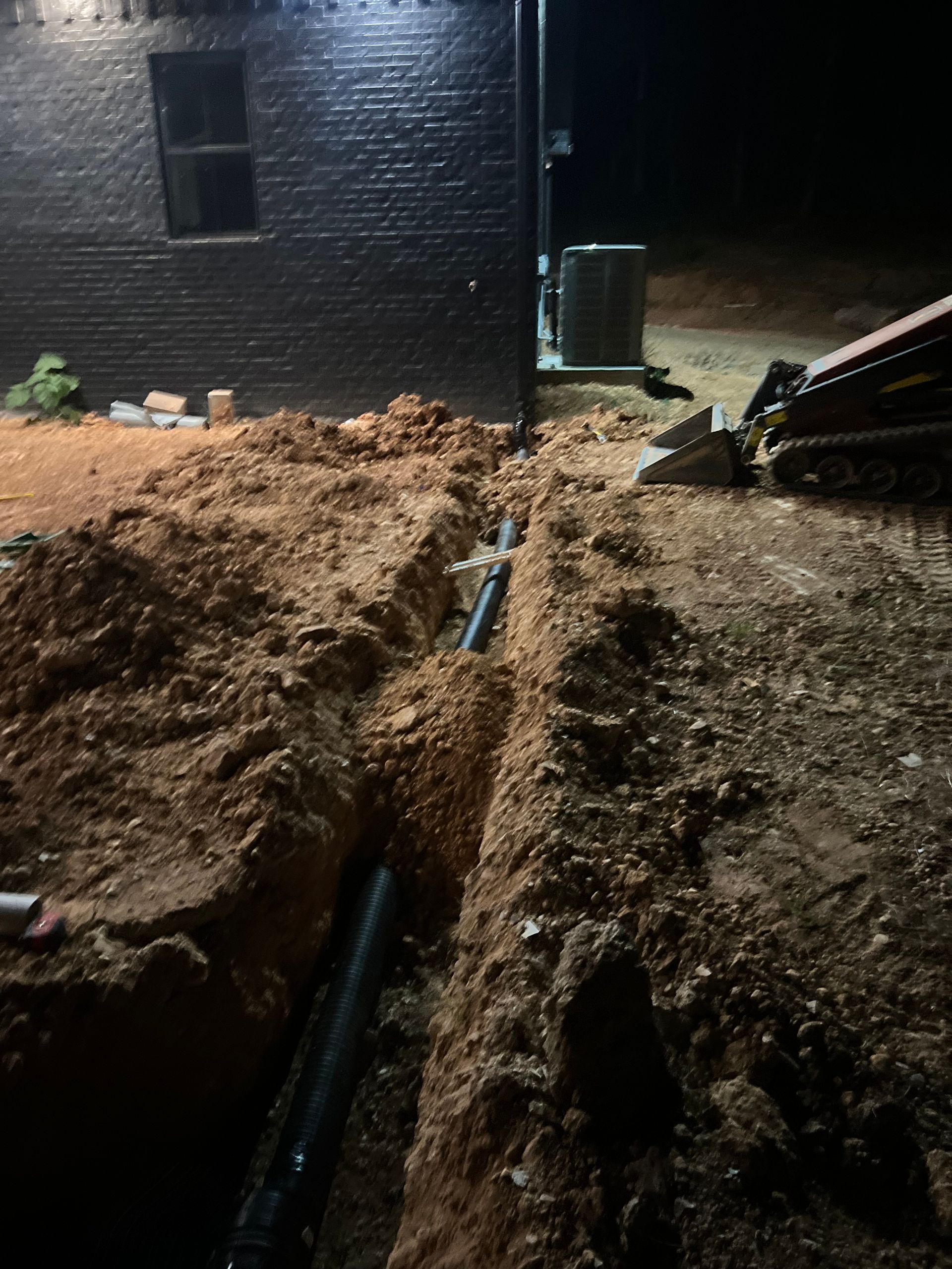 Nighttime trench excavation beside a dark brick building, with dirt piles and exposed black pipes.