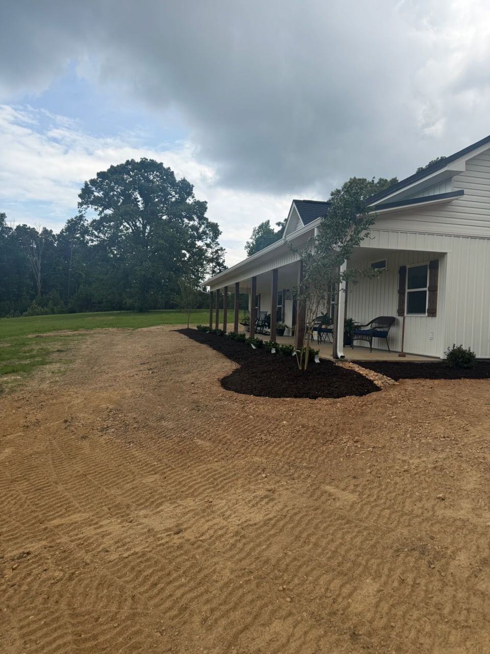 White house with porch on a gravel lot beside a grassy field under cloudy sky