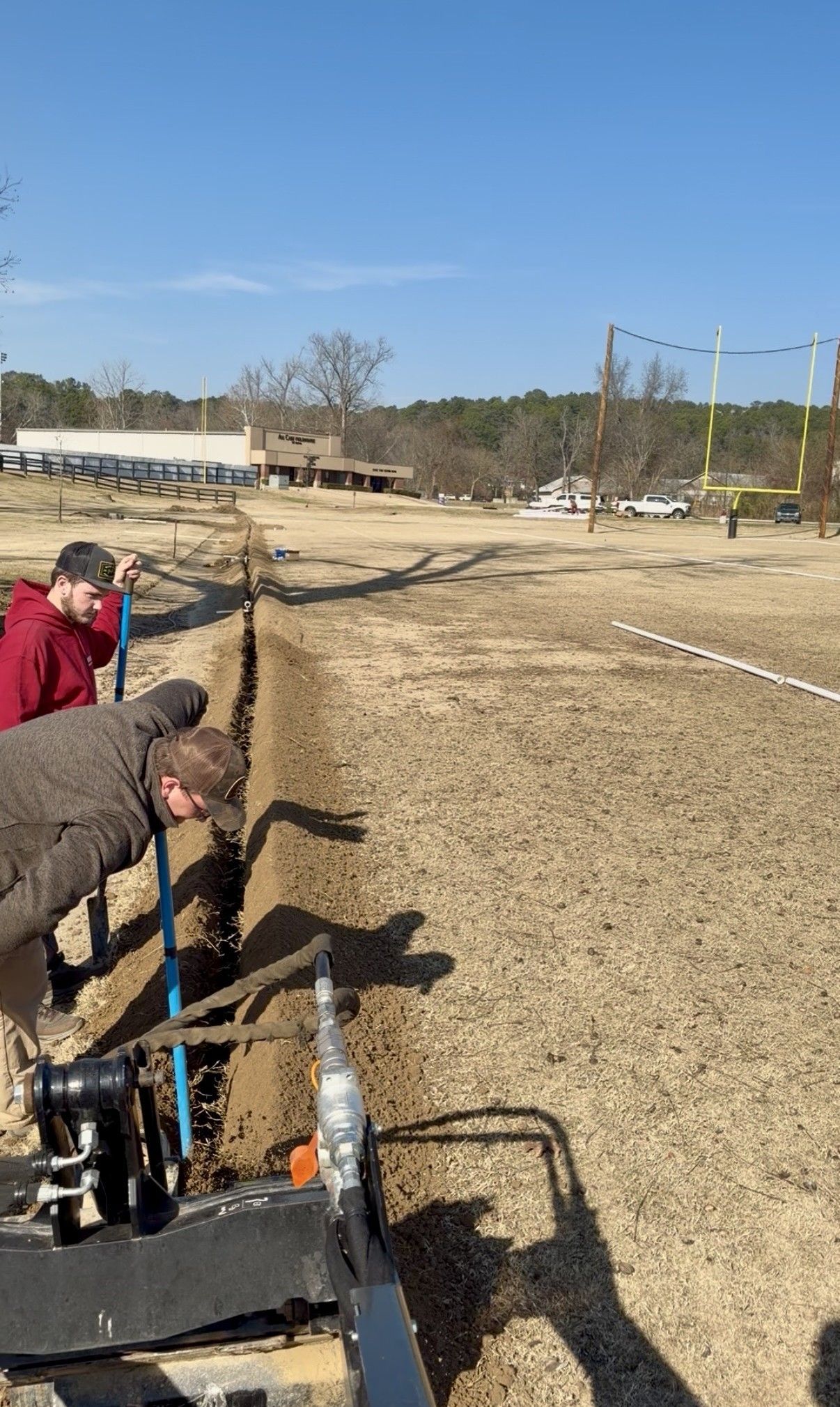 Workers using a trenching machine beside a long dirt trench on a sunny construction site