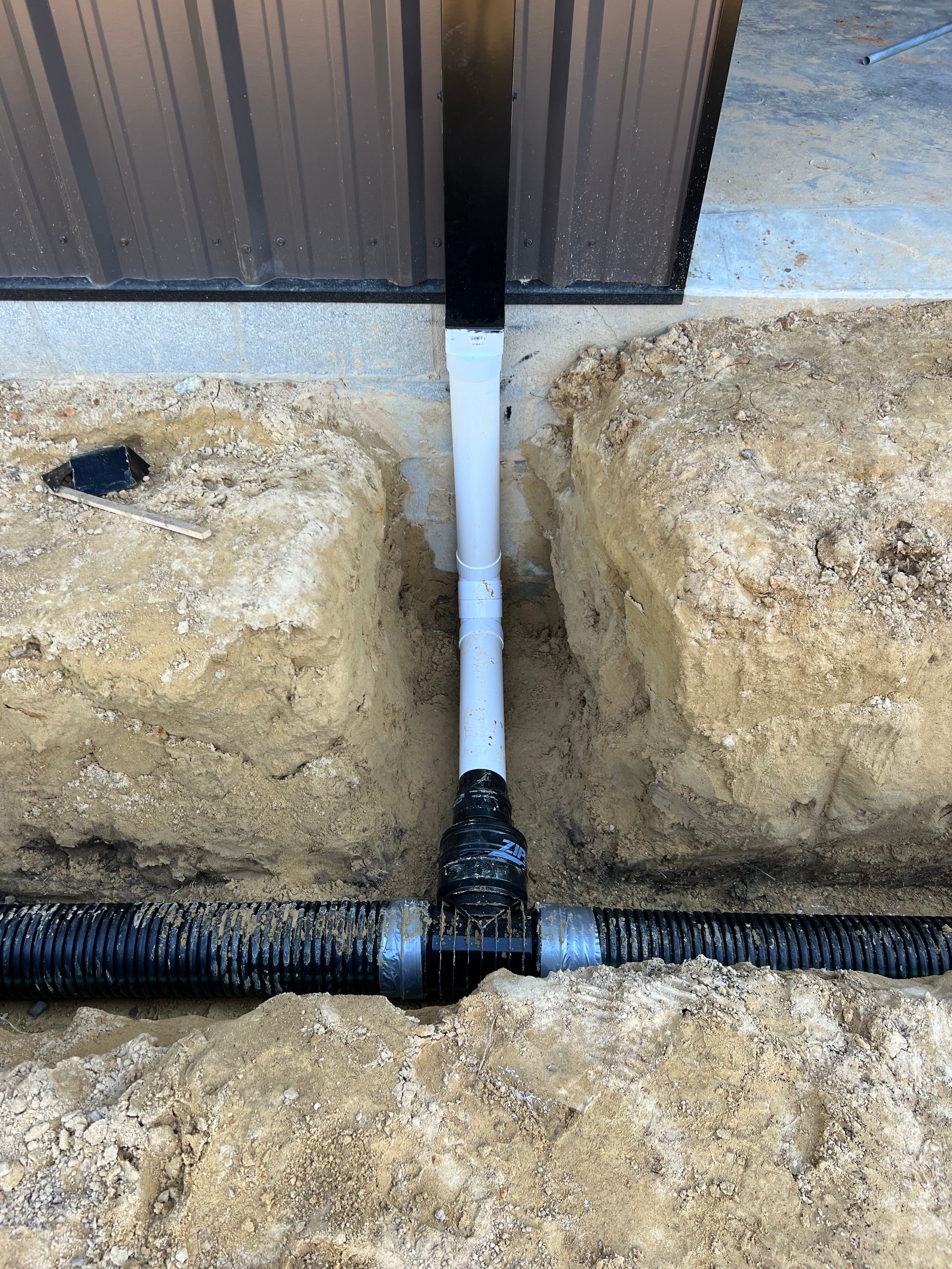 White PVC pipe and black drain line meeting between two large beige rocks beside a wall.